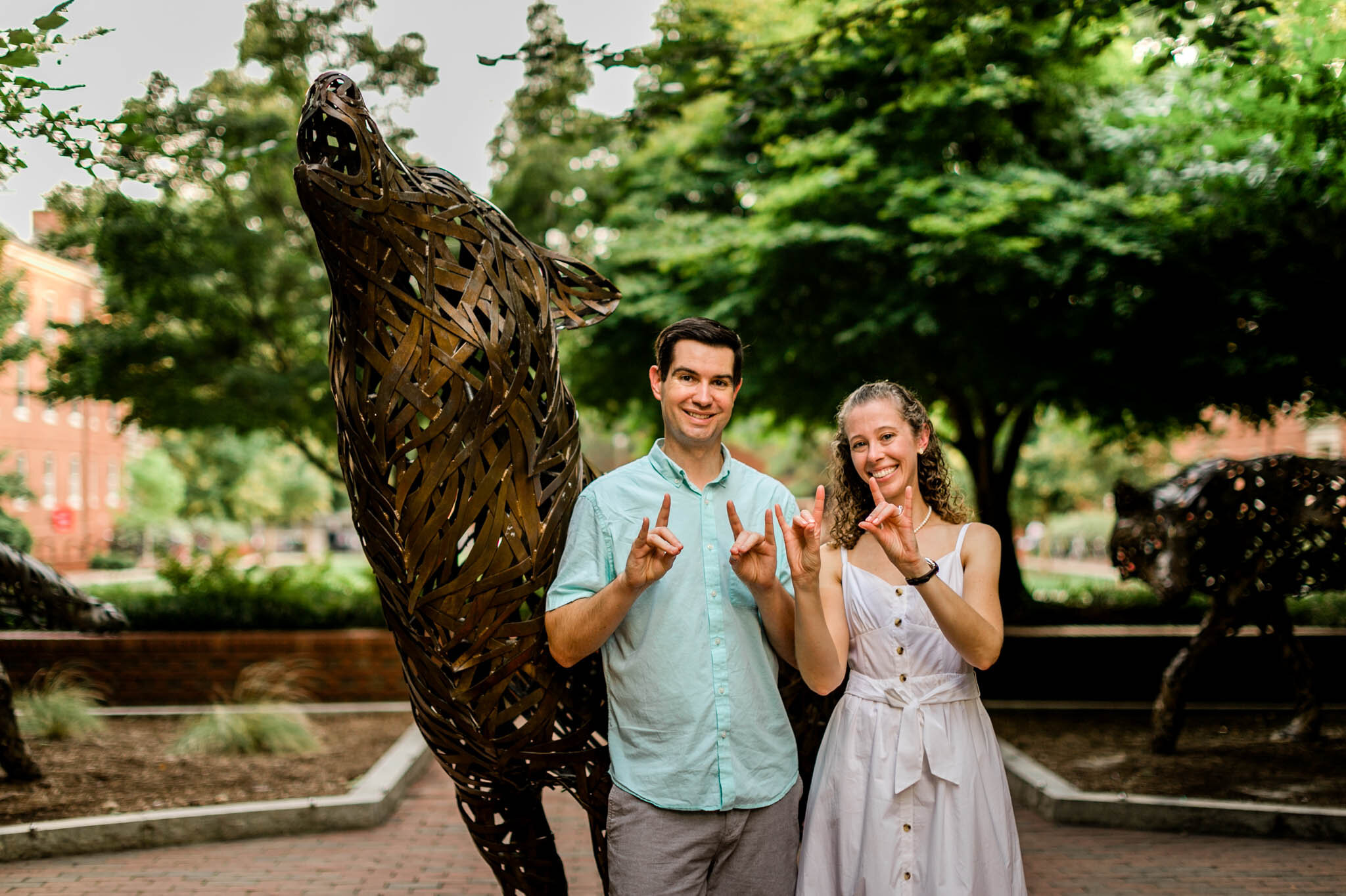 Raleigh Engagement Photographer | NC State University | By G. Lin Photography | Couple making wolfpack signs and standing next to wolf statue