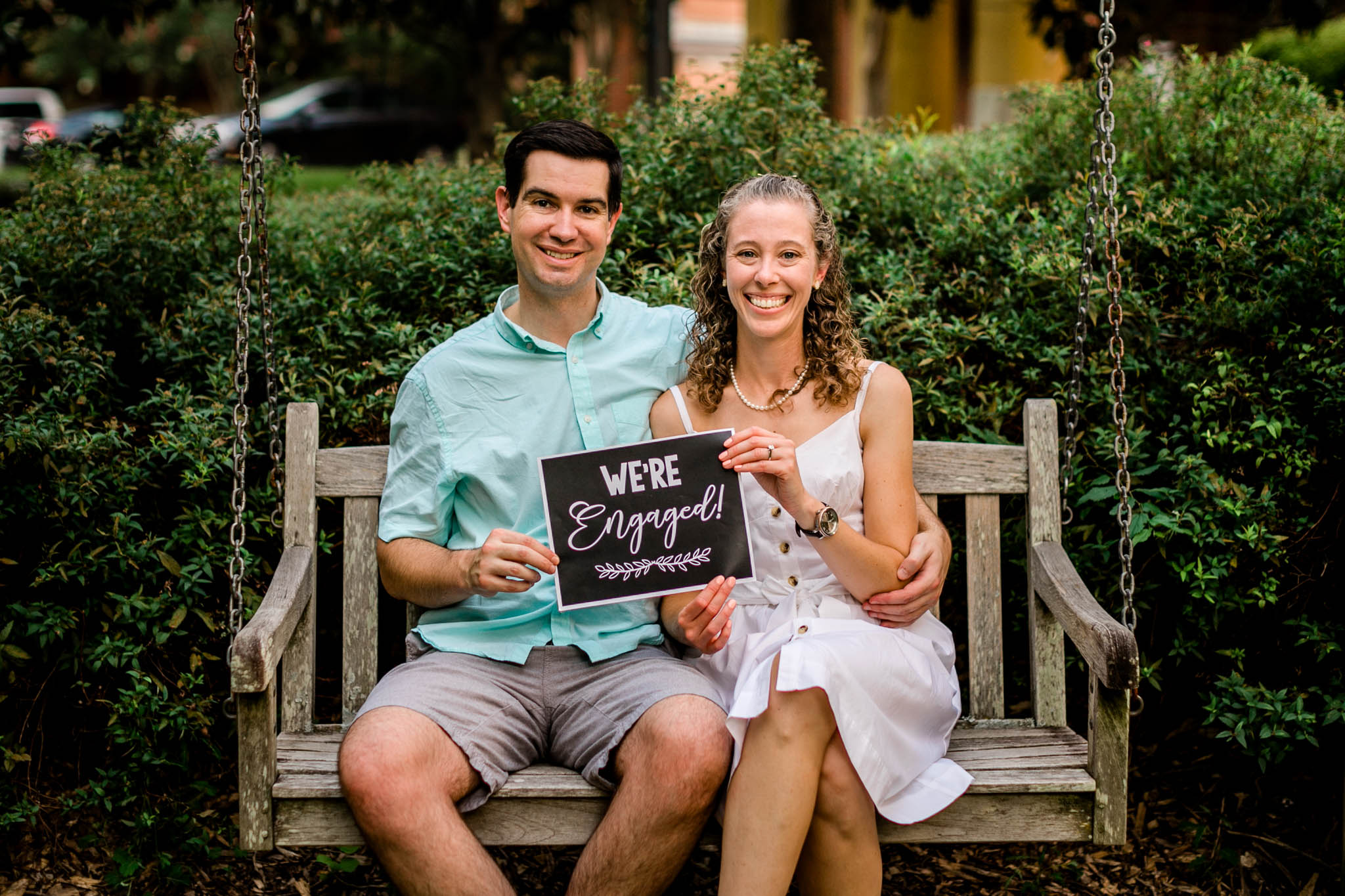 Raleigh Engagement Photographer | NC State University | By G. Lin Photography | Couple holding "we're engaged" sign and sitting on bench