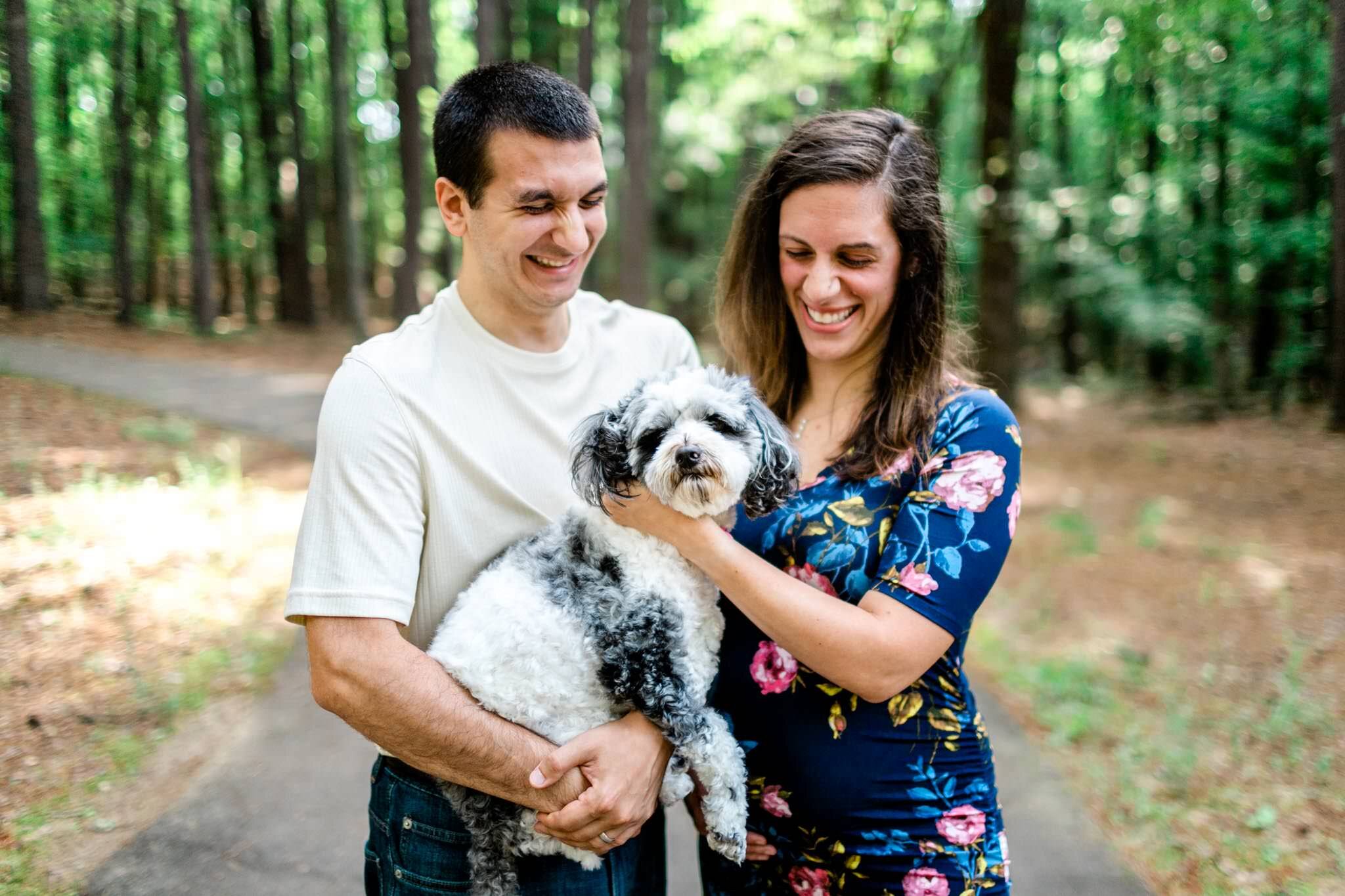 Raleigh Maternity Photographer | By G. Lin Photography | Umstead Park | Couple holding dog in their arms outside in park laughing