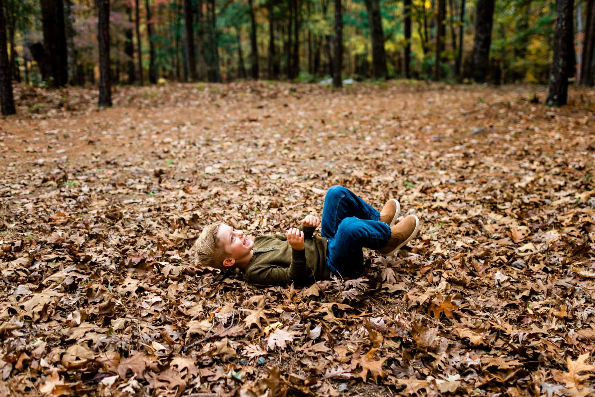 Raleigh Family Photographer at Umstead Park | By G. Lin Photography | Young boy rolling in pile of brown leaves
