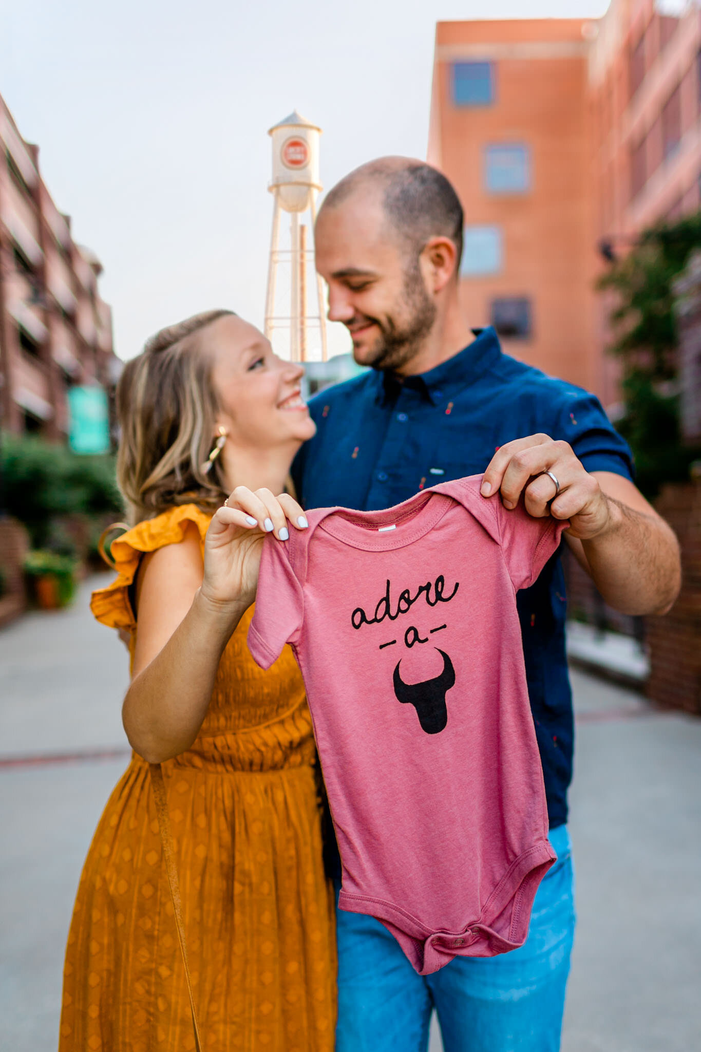 Durham Maternity Photographer | By G. Lin Photography | Woman and man holding pink onsie together | American Tobacco Campus