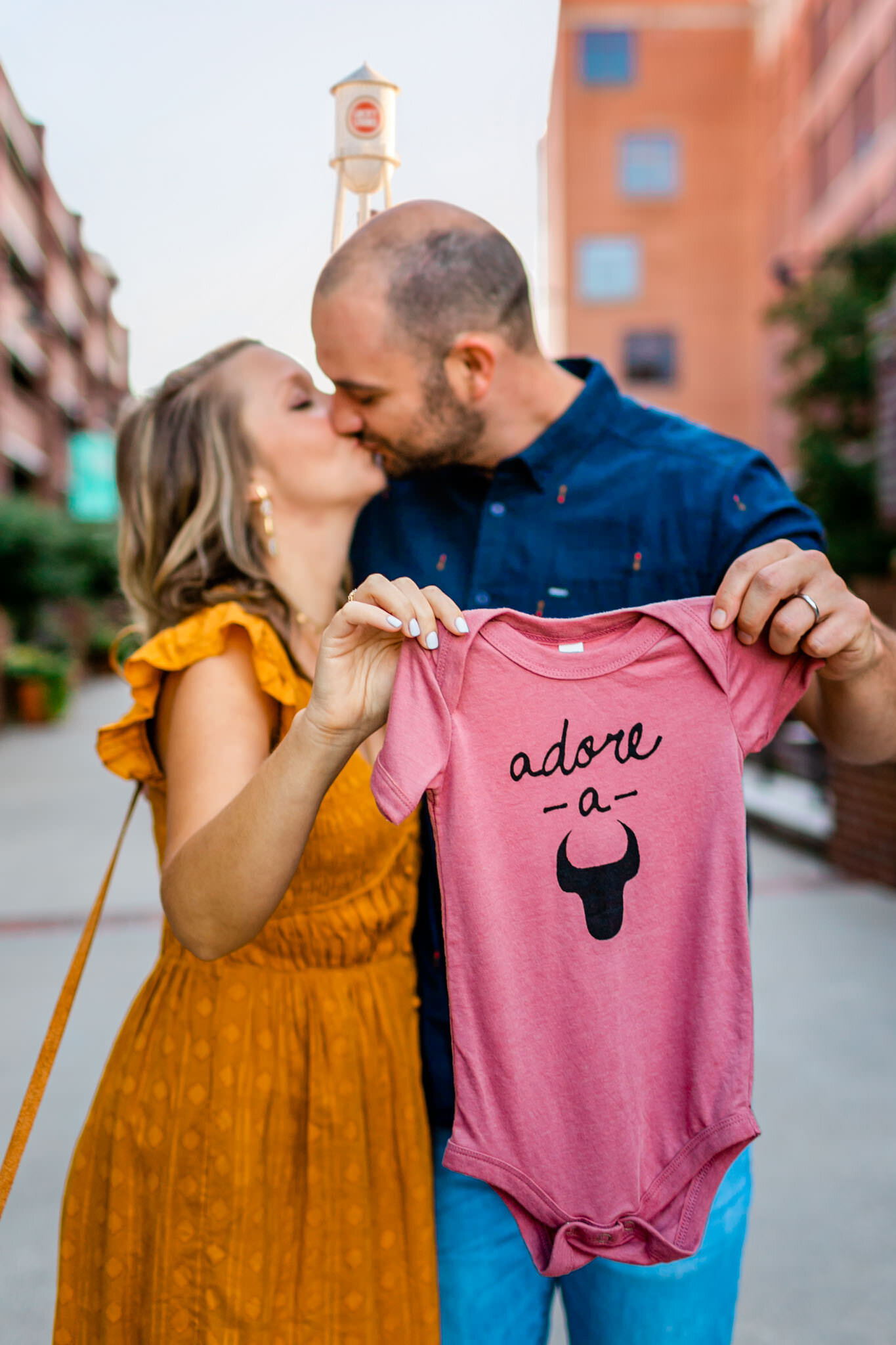 Durham Baby Announcement Photography | By G. Lin Photography | Man and woman kissing and holding pink baby onsie