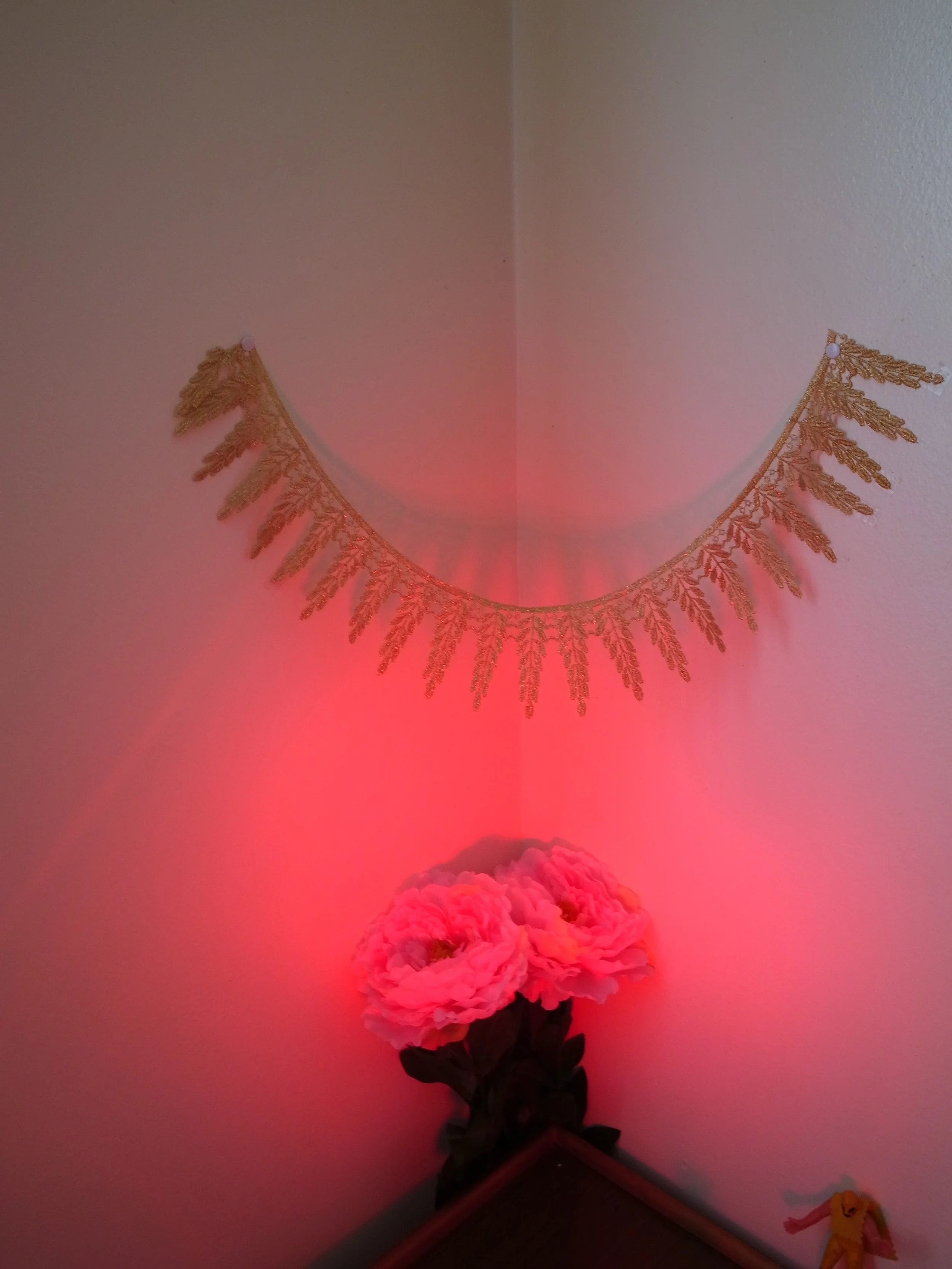 Decorative gold fringe banner hung on a wall above pink floral arrangement illuminated by pink lighting in a corner of a room.