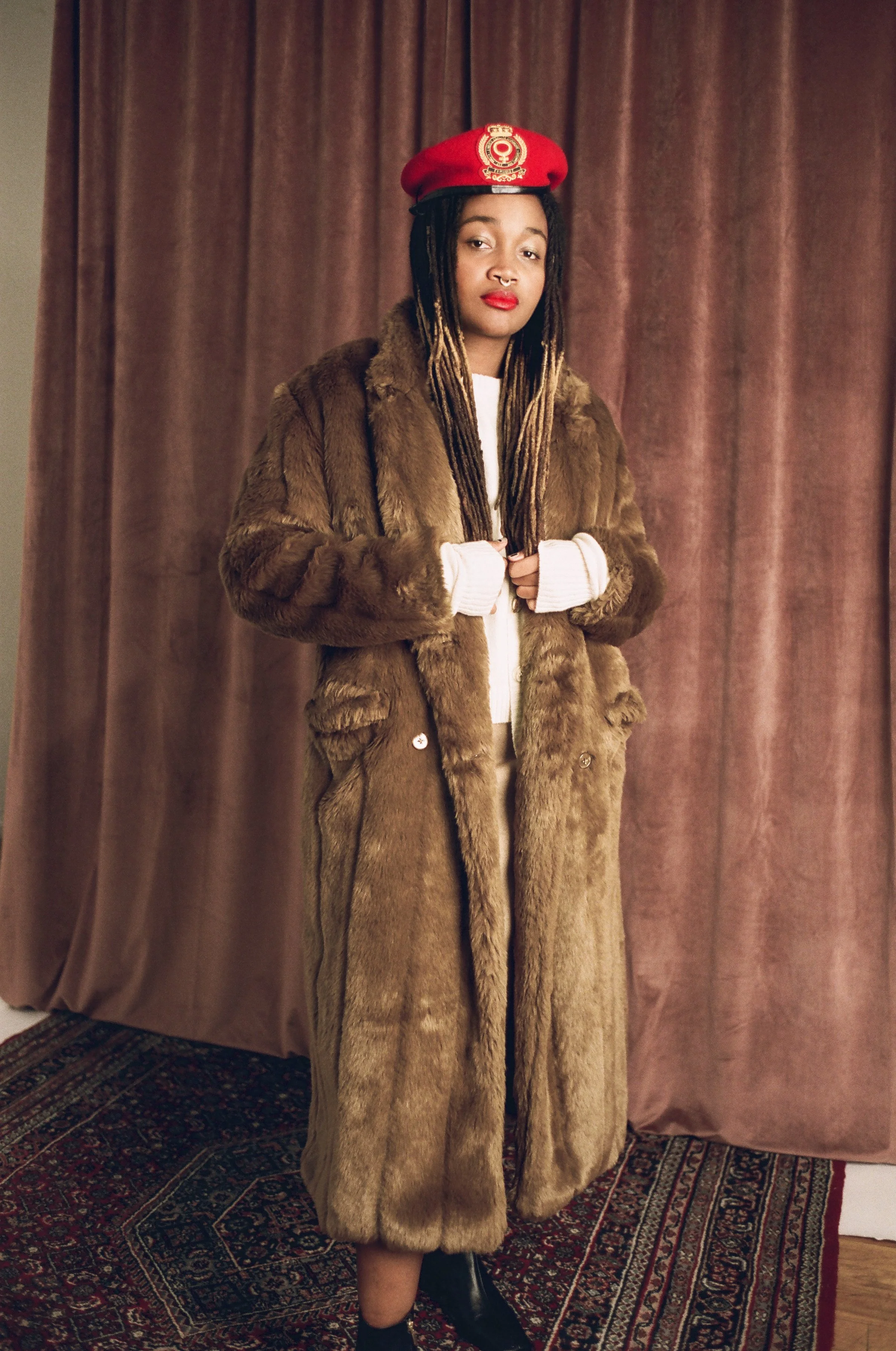 A woman wearing a felt red beret with a gold emblem, a brown fur coat, white sweater, and black boots, standing in front of pink curtains.