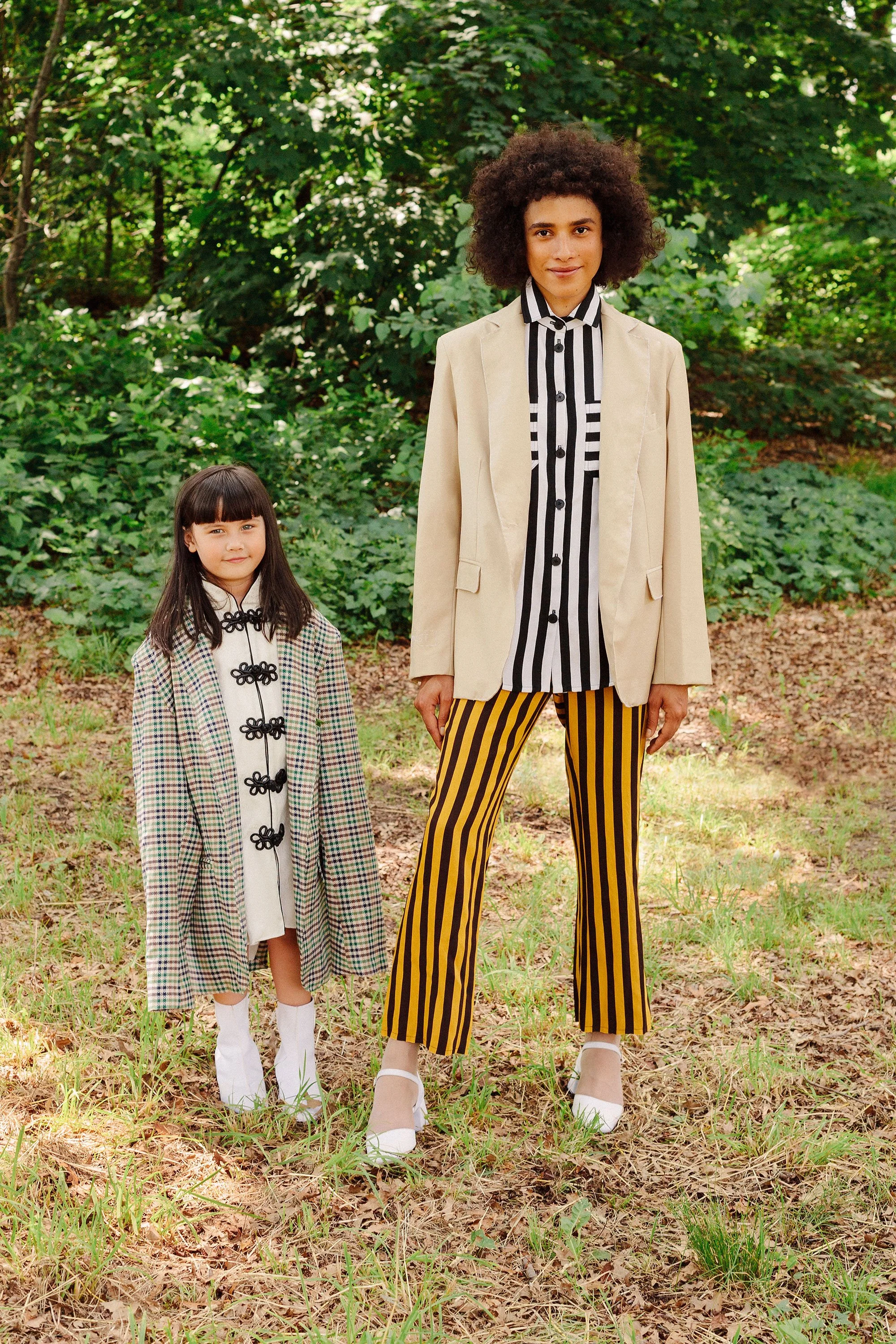 Two women, one young and one older, standing outdoors on a grassy area with trees in the background. They are dressed in fashionable clothing, with the older woman wearing a beige blazer and yellow-and-black striped pants, and the young girl wearing 