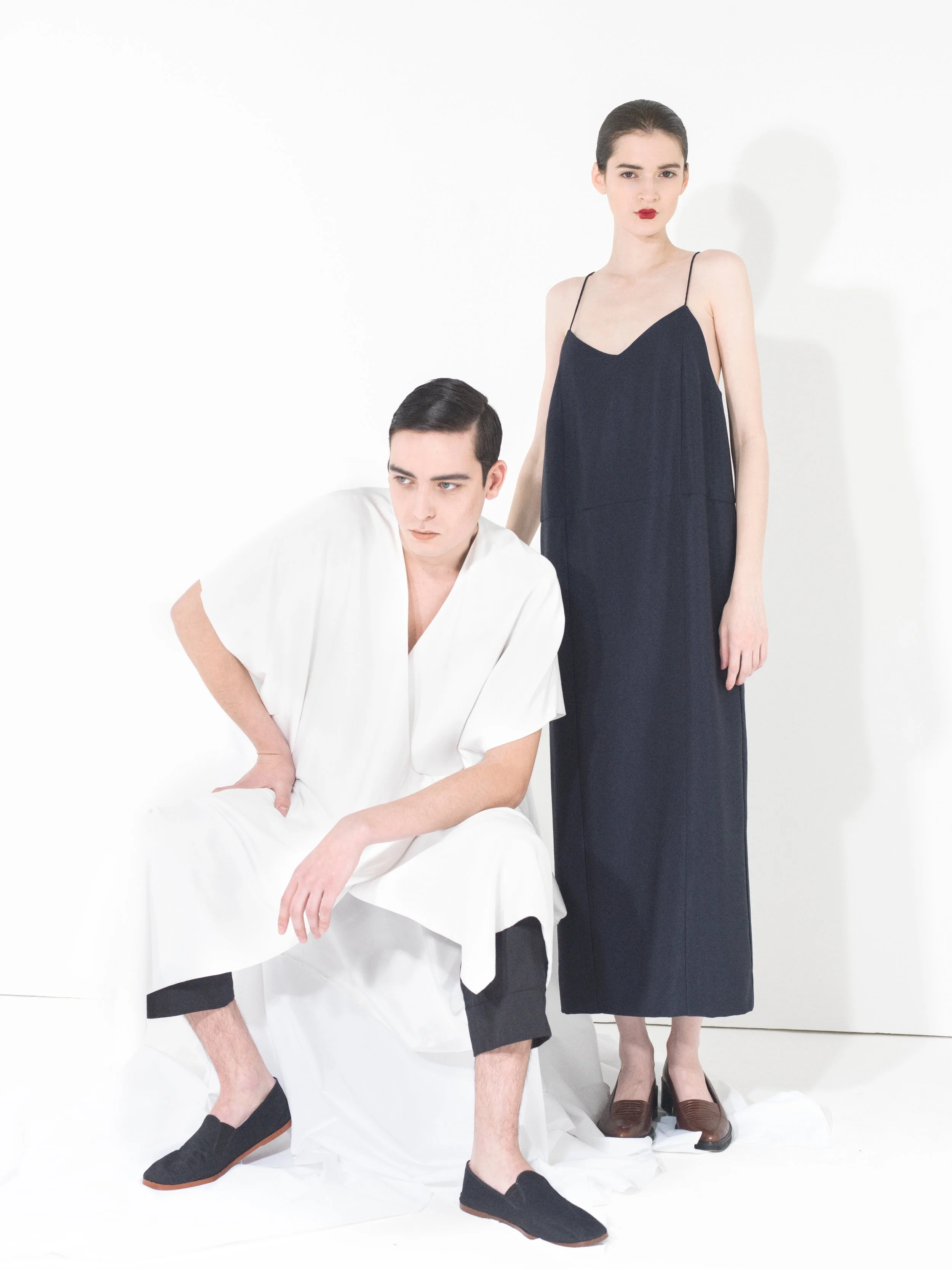 A man and a woman with dark hair and light skin posing against a plain white background. The man is seated, wearing a white robe with black pants and black slip-on shoes. The woman is standing, wearing a long navy blue dress with thin straps and brow