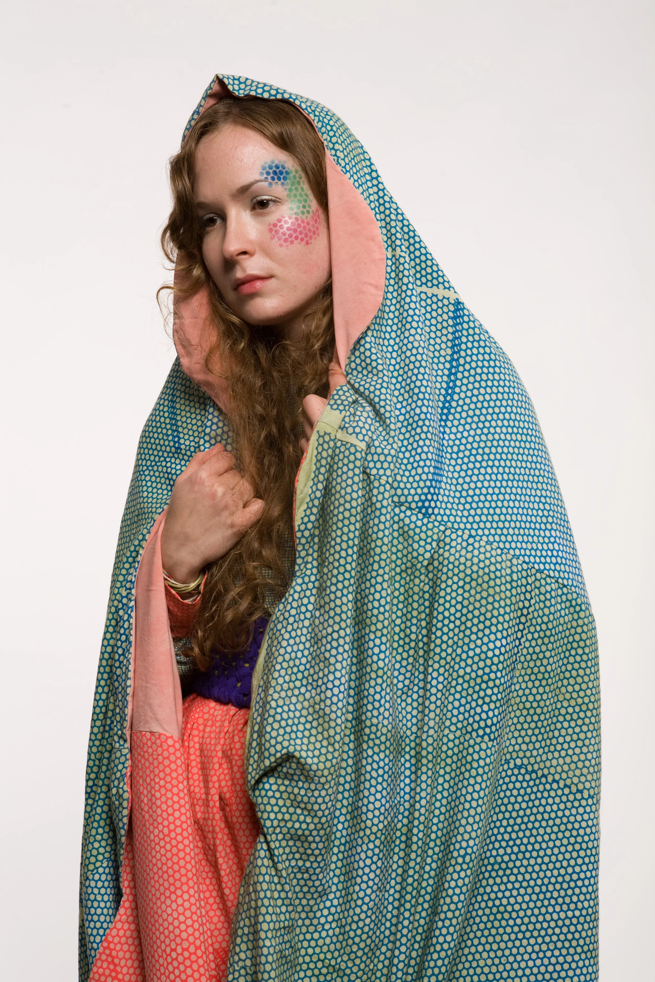 A young woman with long curly hair, wearing a colorful patterned hooded cloak, with her face painted with pink, green, and blue dots.