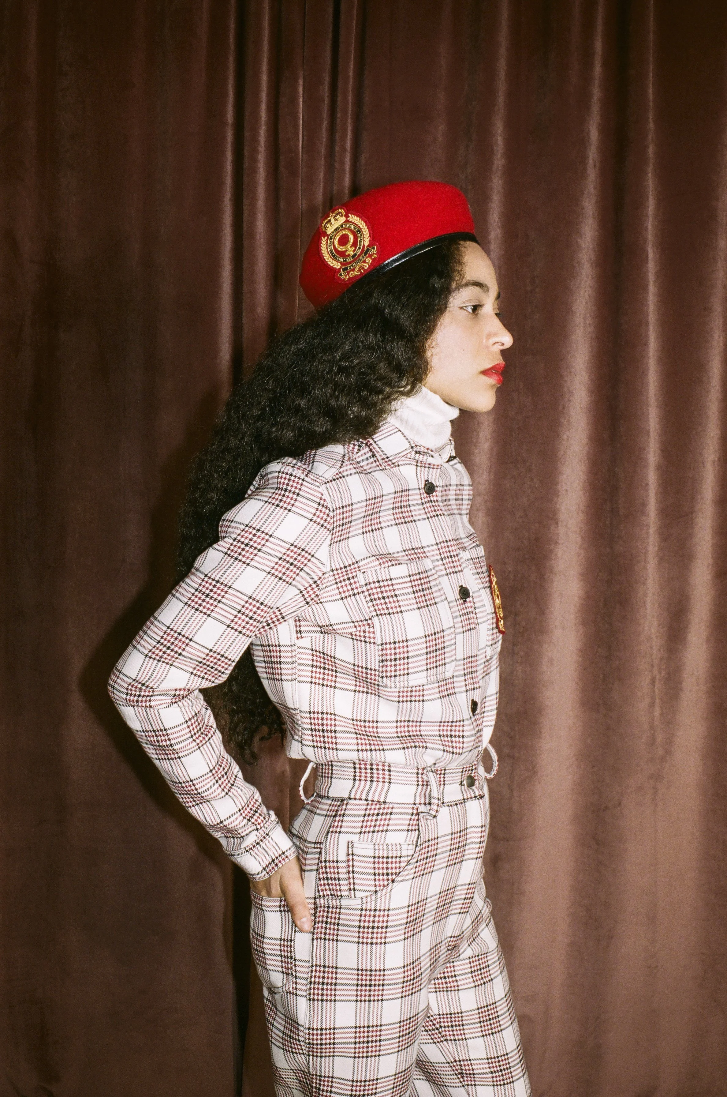 A woman with long curly hair dressed in a plaid shirt and pants with a red hat standing against a brown curtain background.