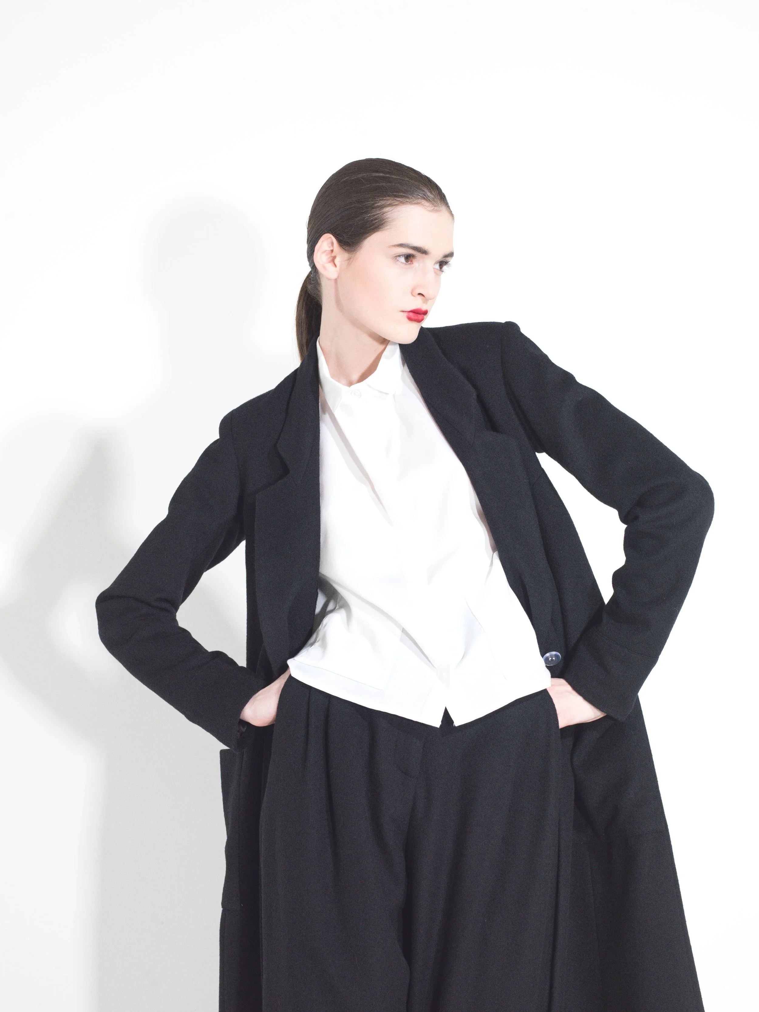 Woman in business attire with hands on hips, wearing a black blazer, white shirt, and black trousers, standing against a white background.