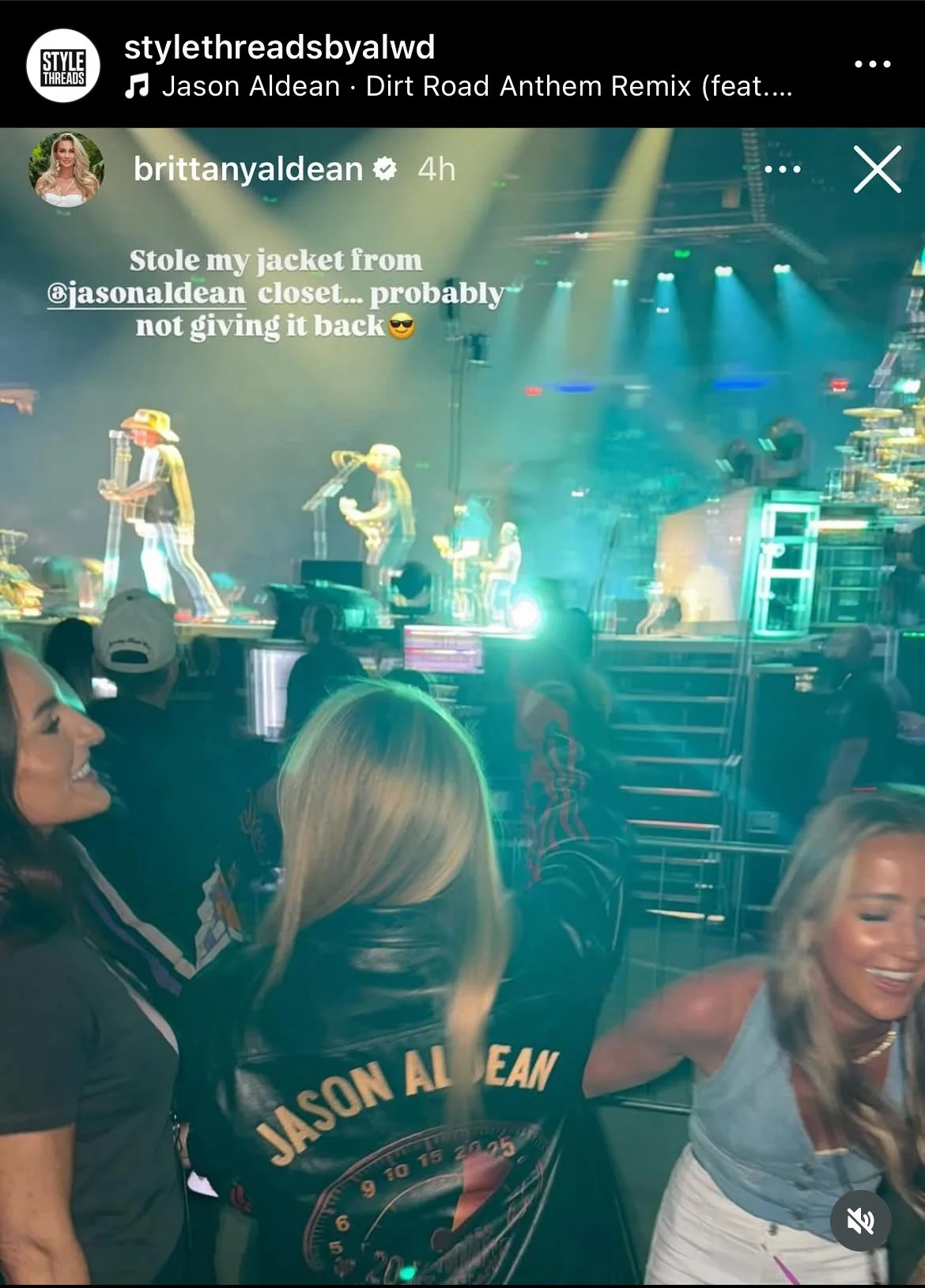 Two women in the foreground are smiling and talking at a concert or show with a stage in the background. The woman on the left is wearing a black jacket with Jason Aldean's name and logo on the back, and the woman on the right has blonde hair, is wearing a grey top and white pants, and is holding a drink. The stage has performers, bright stage lights, and equipment, with an audience visible in front.