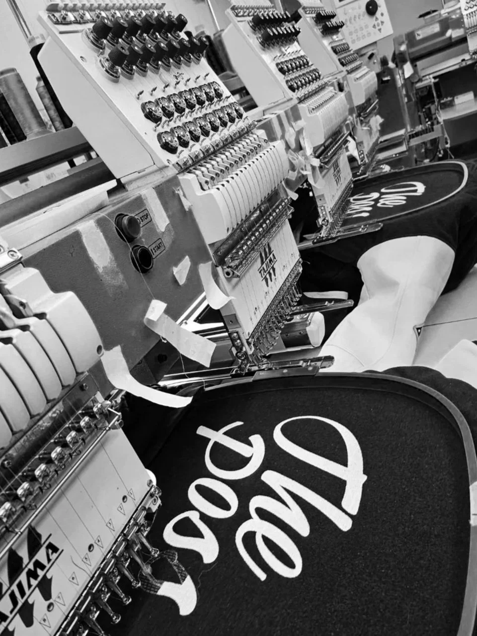 Close-up of an industrial embroidery machine with threads and controls, stitching a design onto a fabric. There is a person in a dark hoodie with white text on the fabric.