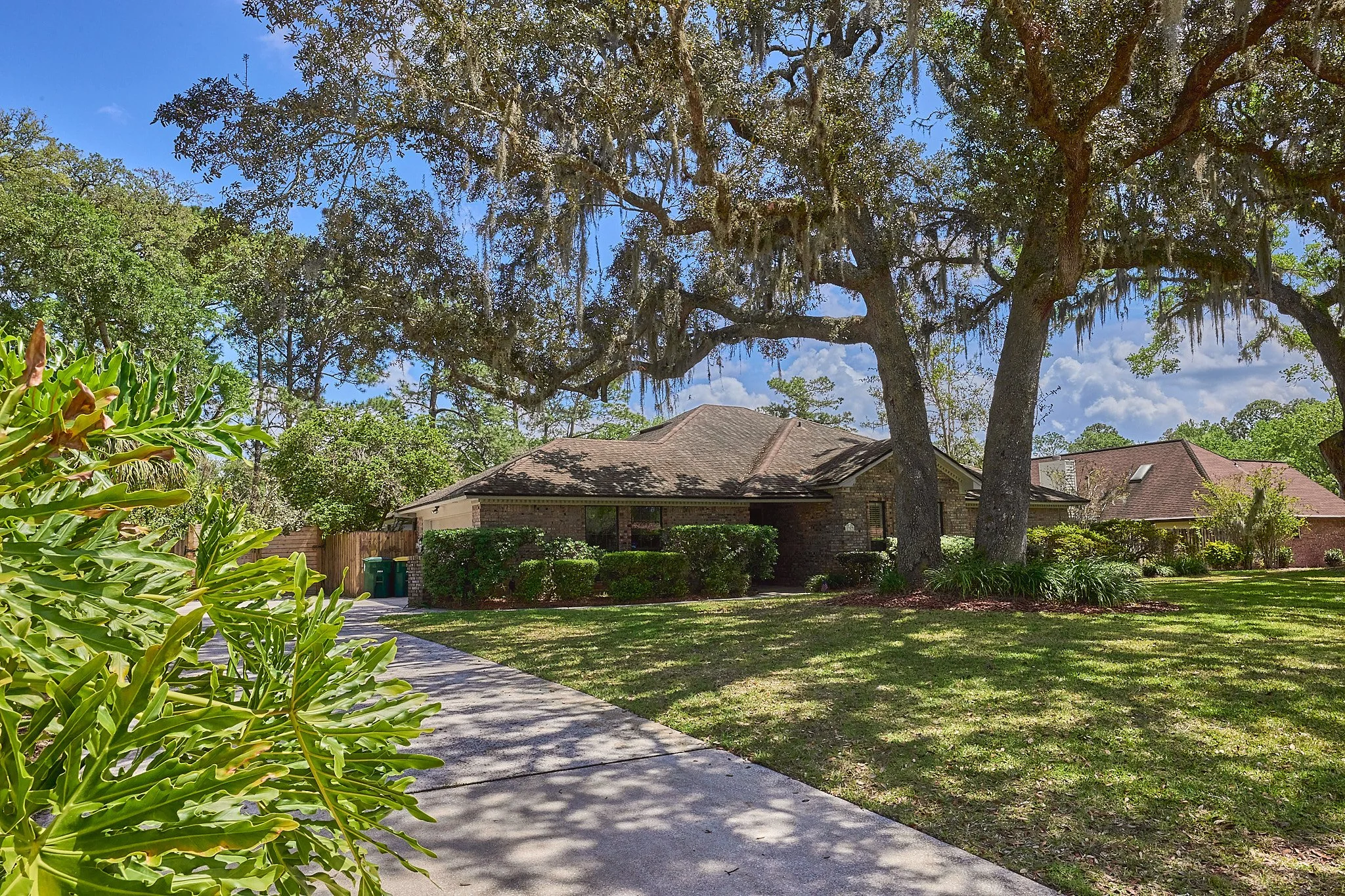 Residential house with a front yard, large trees, and a concrete walkway leading to the house in a suburban neighborhood on a sunny day.