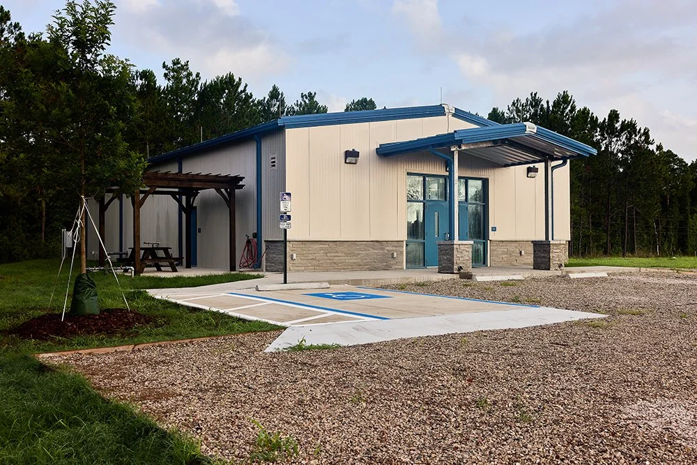 A modern building with beige and blue exterior, a small covered entrance, parking spaces including a designated accessible spot, surrounded by trees and a partly cloudy sky.