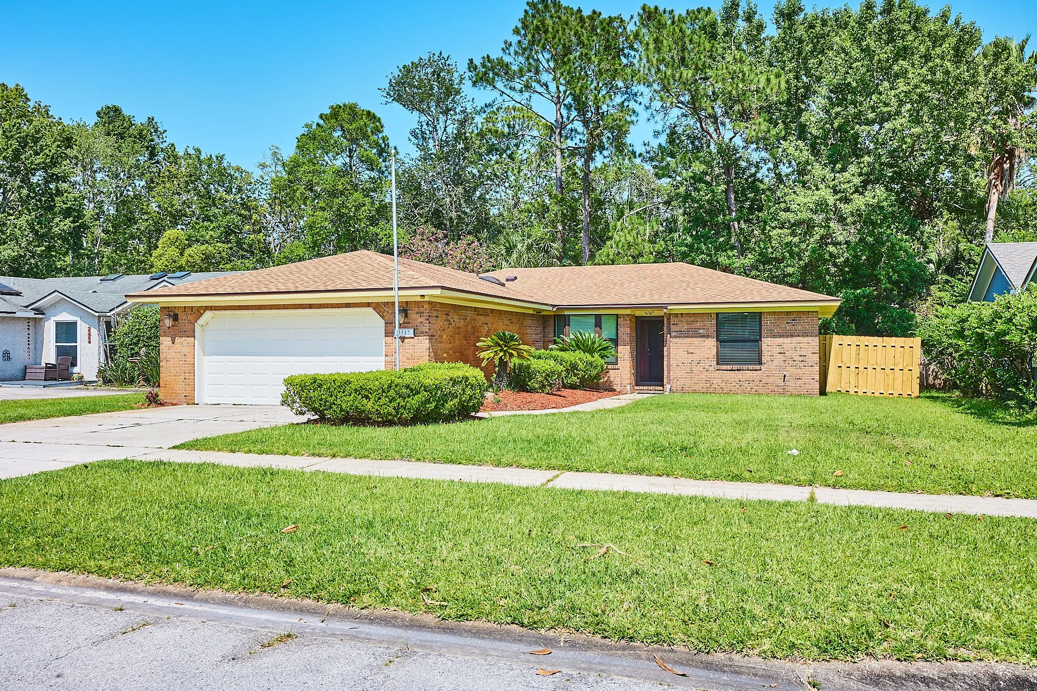 A single-story brick house with a brown shingle roof, white garage door, and a small front shrubbery. The house is surrounded by a green lawn and established trees, with a driveway leading to the garage and a sidewalk in front.