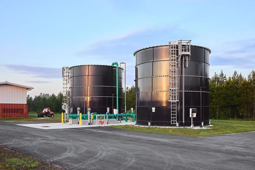 Two large black cylindrical storage tanks with metal ladders and green piping, situated outdoors on a paved area with a small building and trees in the background.