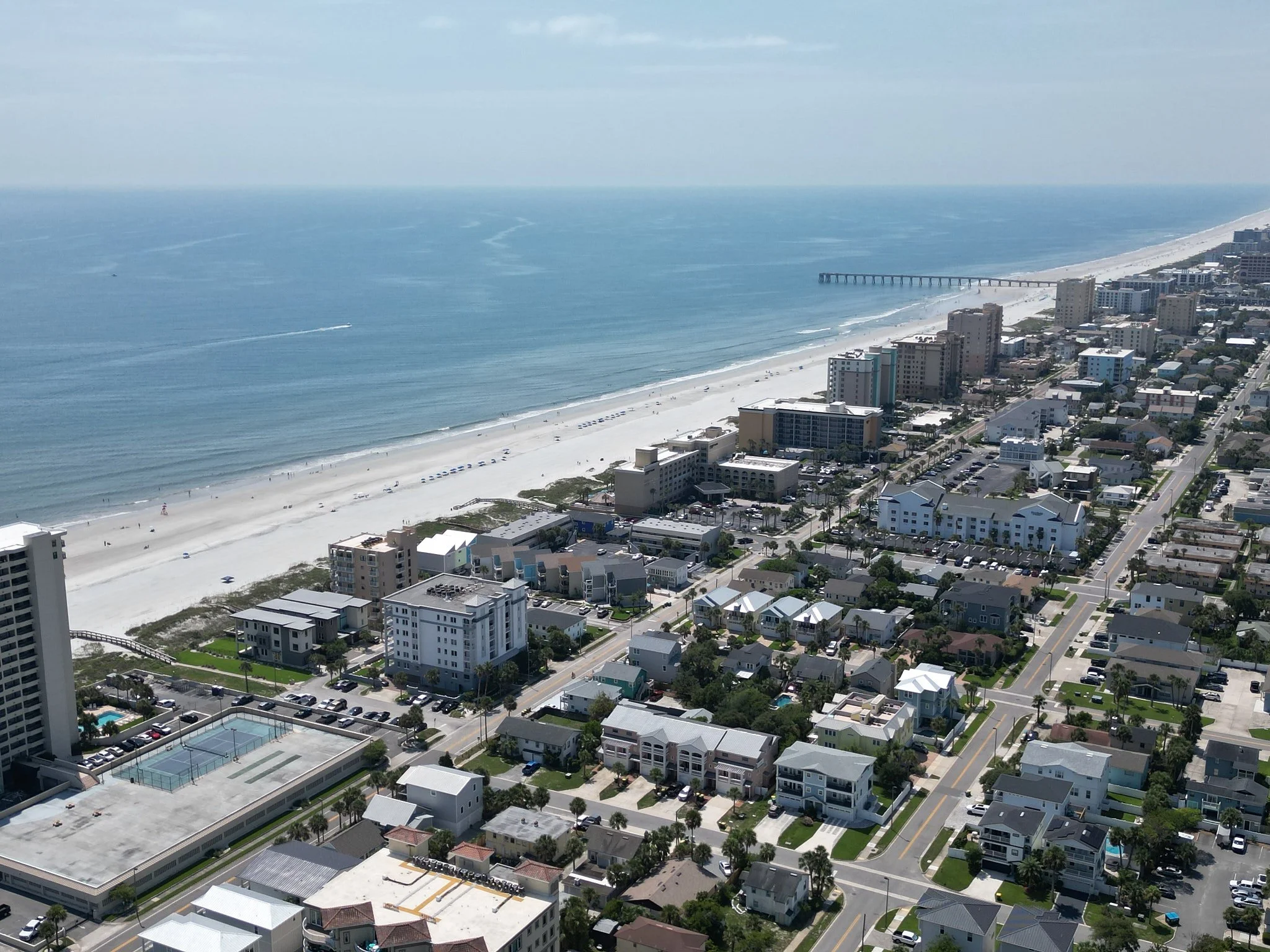 Aerial view of a beachside city with high-rise buildings, houses, and a long sandy beach along the coastline. A pier extends into the ocean, and a boat leaves a trail in the water.