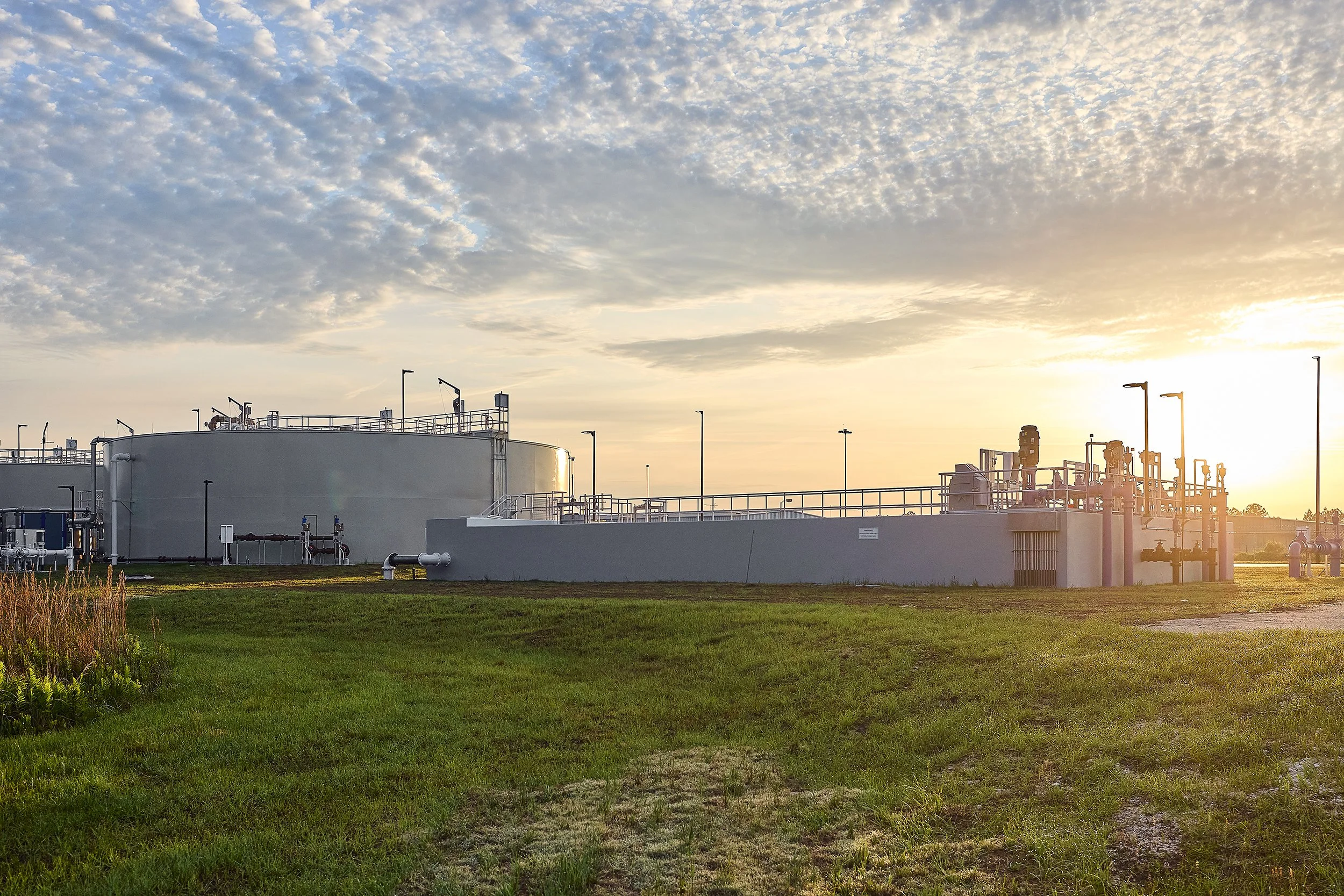 Industrial tanks and piping at a facility during sunset with grassy area in foreground.