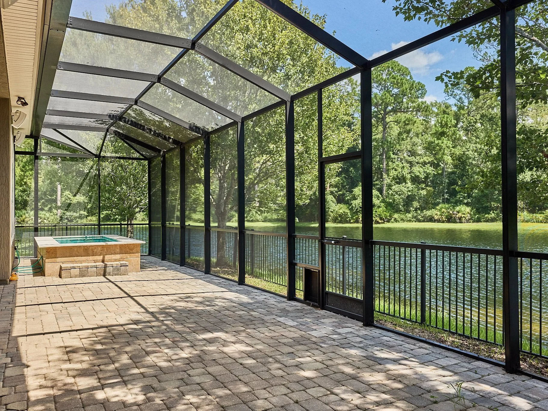 Screened-in patio with a hot tub, brick pavers, overlooking a lake with trees and green foliage.