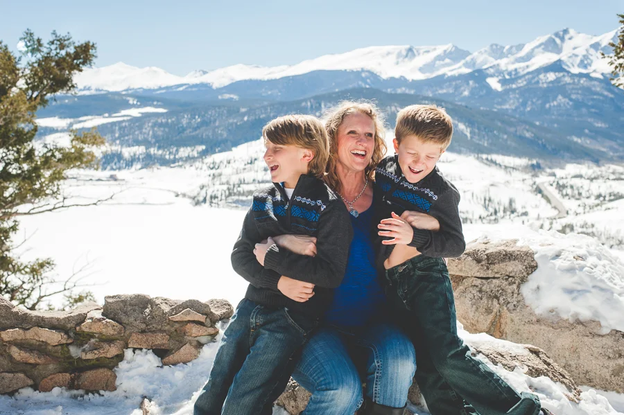 Tickles and giggles break out during a winter family photo shoot in the mountains, overlooking Peak One and Lake Dillon near Breckenridge, Colorado. Mom and two sons.
