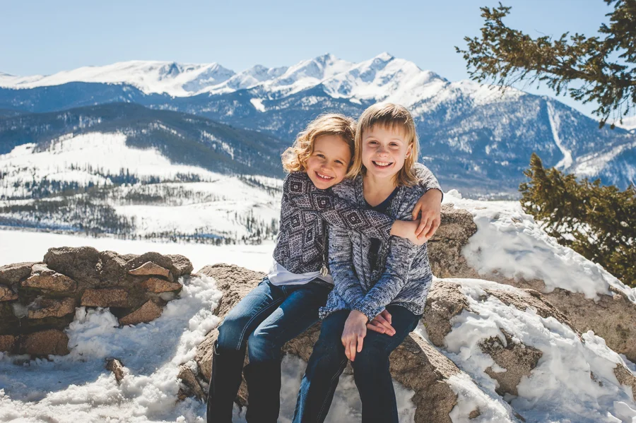 Younger sister hugs older sister during a winter photo shoot in the mountains near Breckenridge, Colorado. Peak One and Lake Dillon are in the background. 