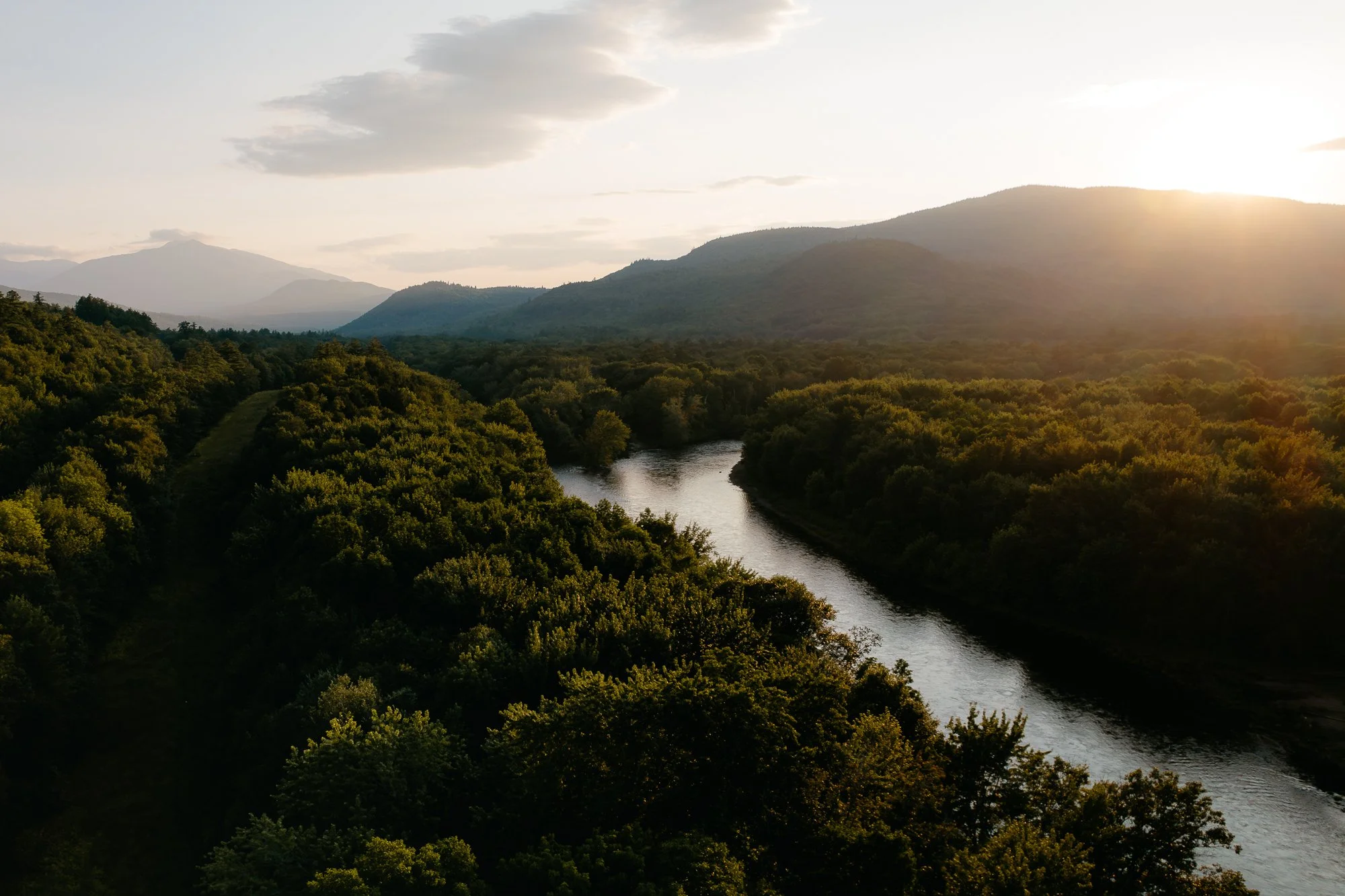 A river flowing through a forested valley with mountains in the background during sunset.