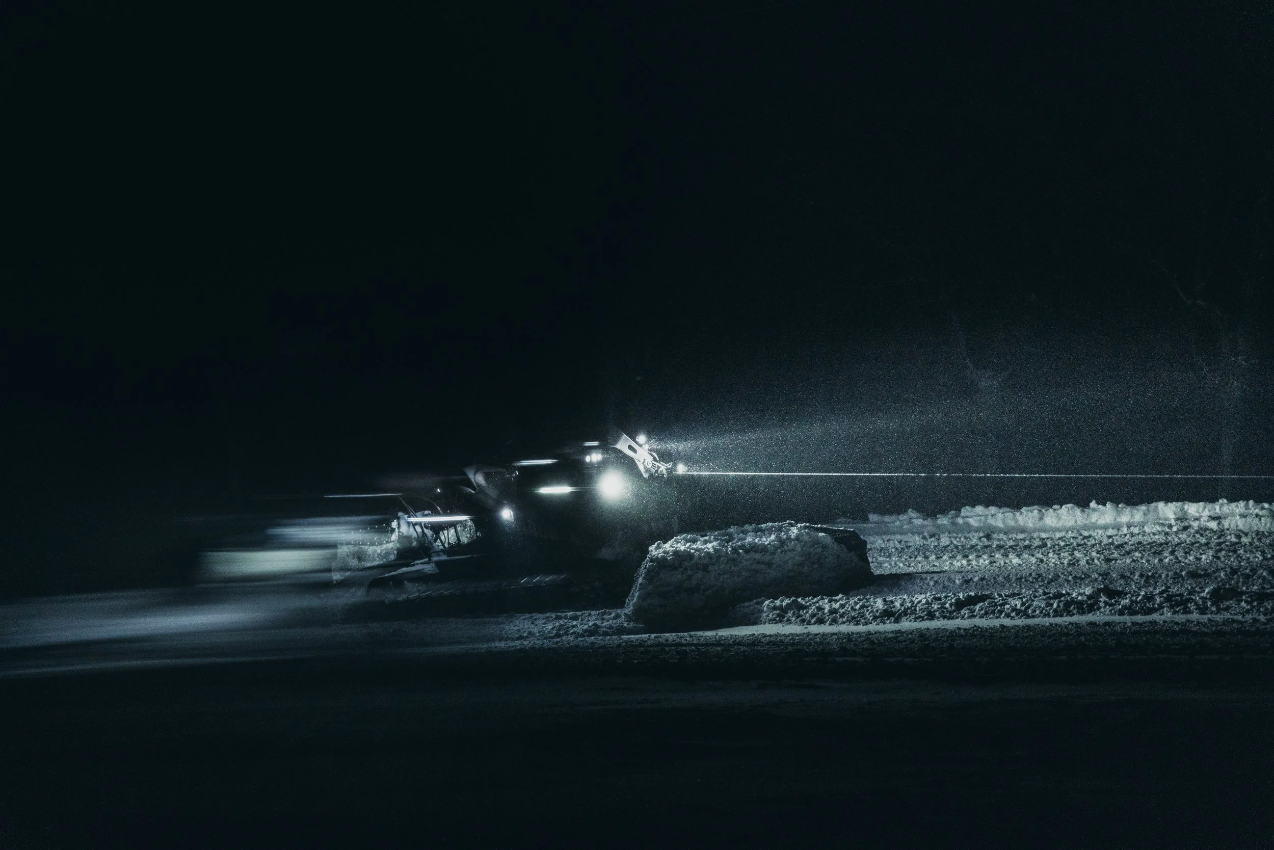 A snowcat vehicle moving through a snowy terrain at night with headlights illuminating the snow.