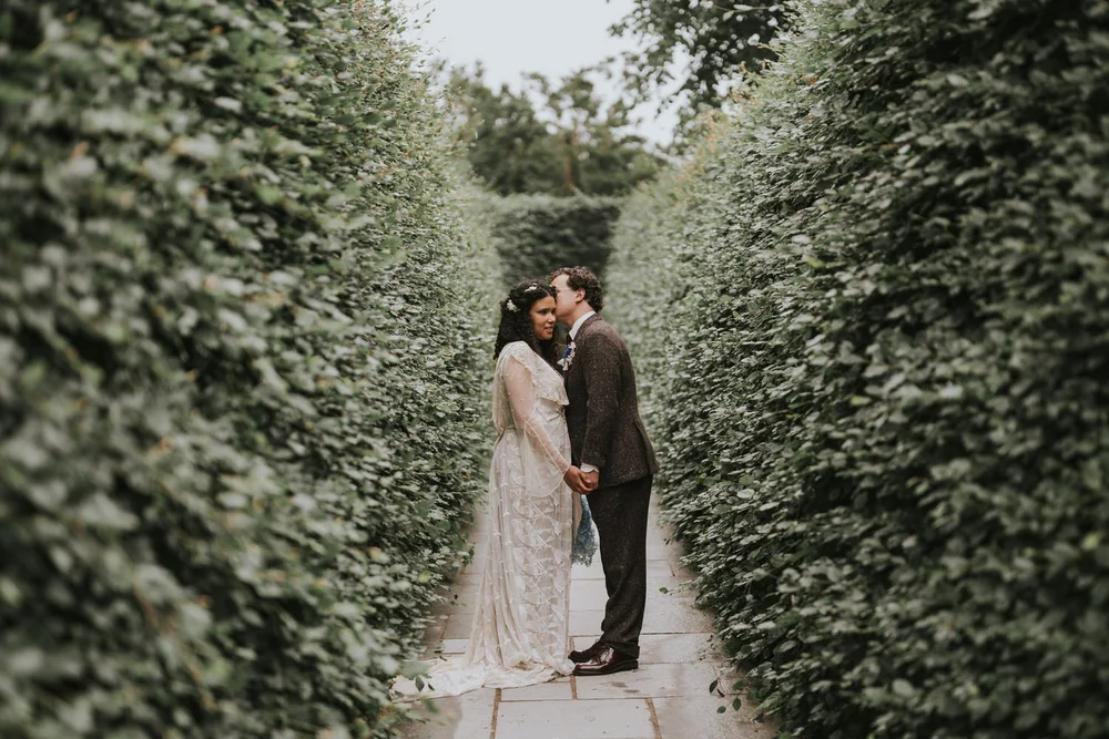 Nneka and Jonathan kissing between the tall hedges on the garden path at Old Court