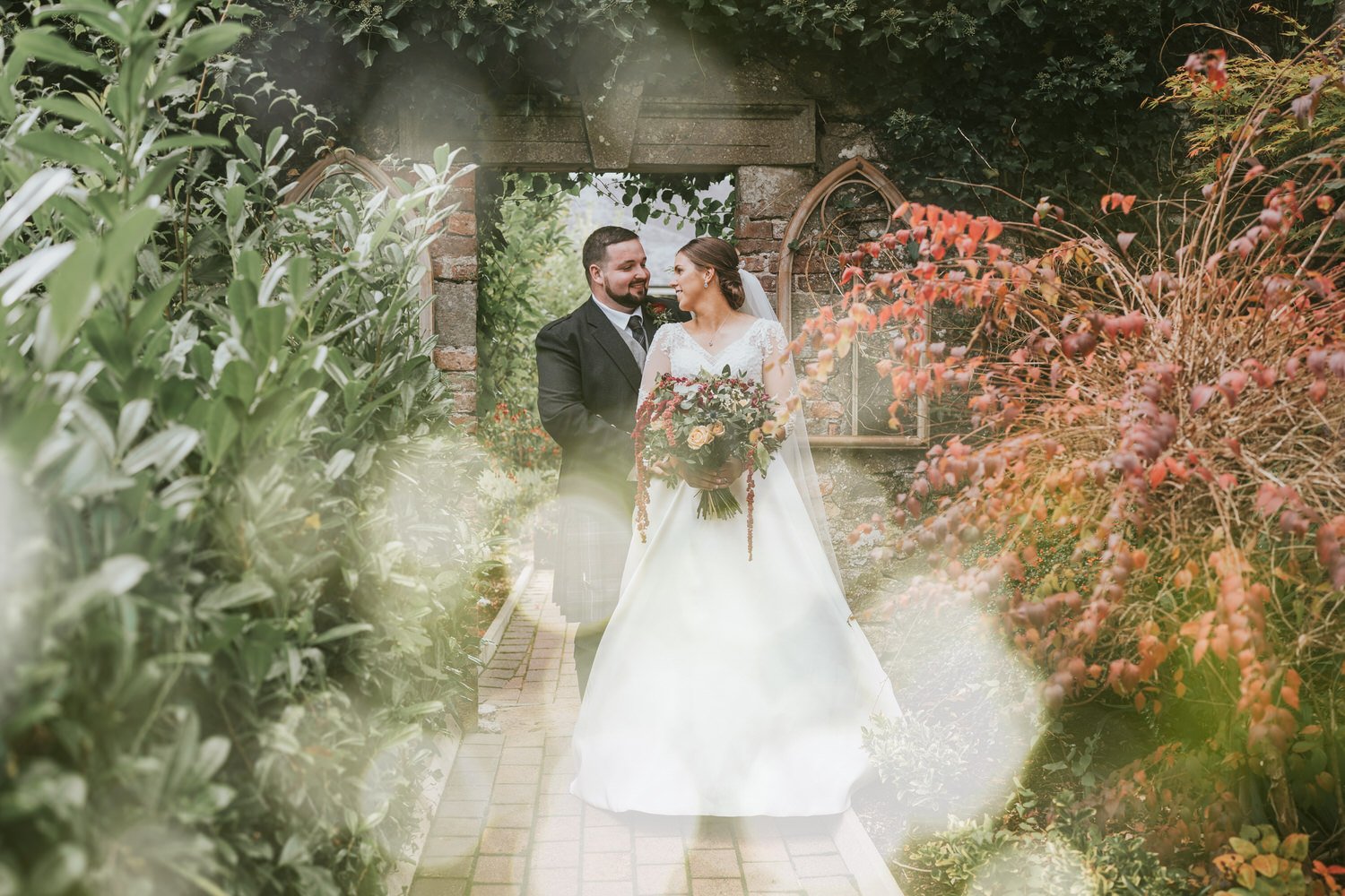 Bride and groom standing together on a brick path in the garden at Corick House, with autumn coloured foliage and a stone archway behind them