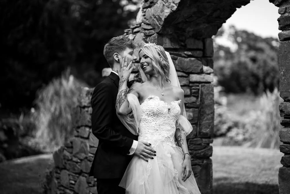 Bride and groom embracing beside an old stone archway in the gardens at Millbrook Lodge, in black and white