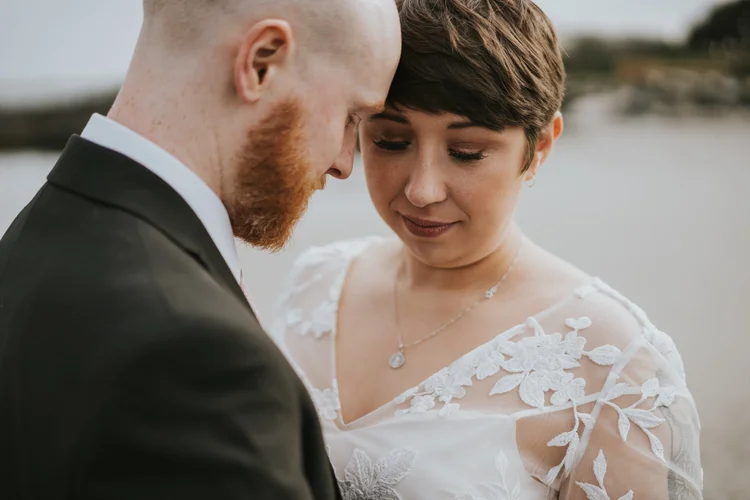 Bride and groom resting their heads together in a quiet moment by the sea on their wedding day at home