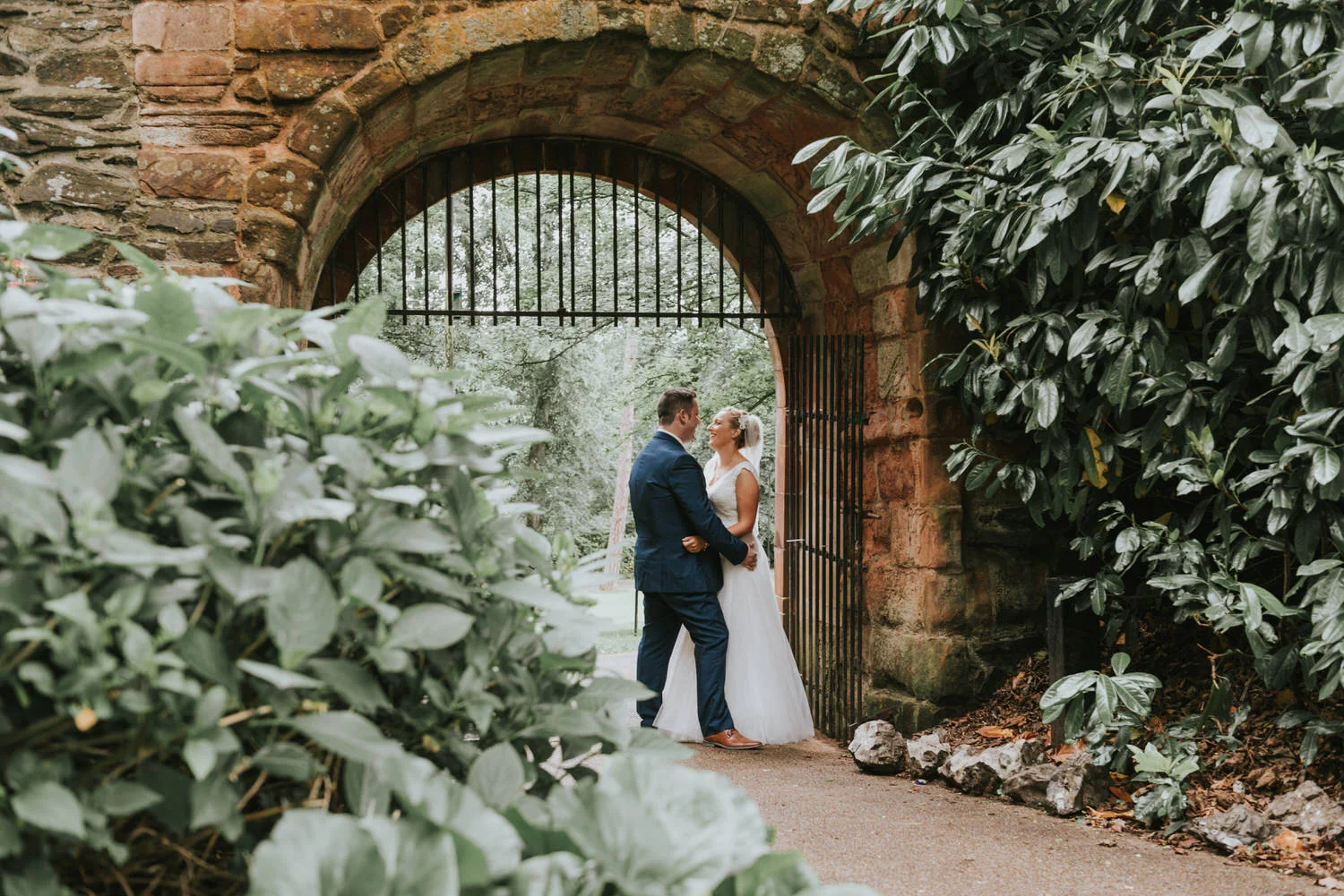 Bride and groom embracing in a stone arched gateway with iron gates surrounded by leafy shrubs at Lagan Valley Island