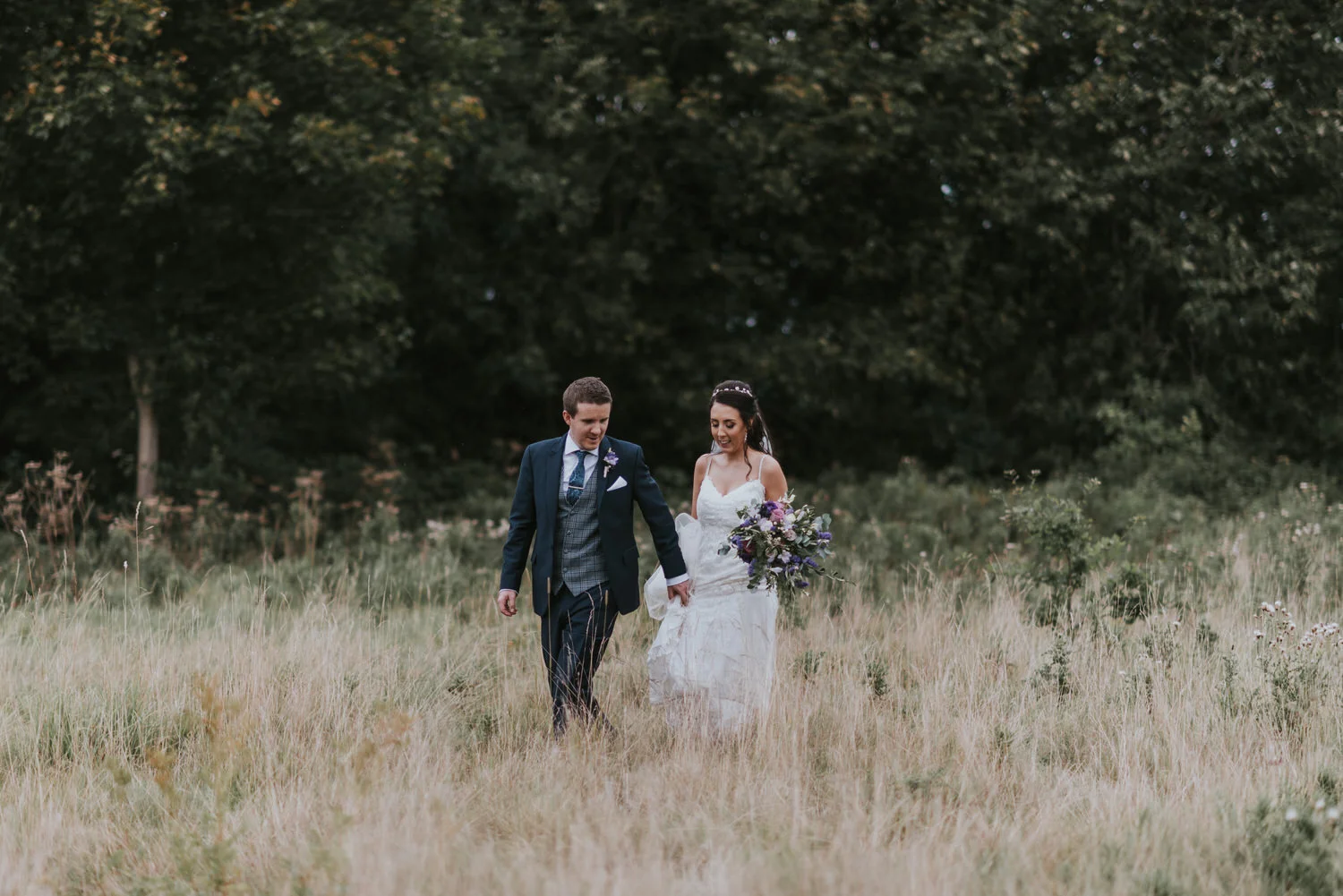 Bride and groom walking hand in hand through long grass at the Kingfisher Country Estate, formerly the Hilton Templepatrick