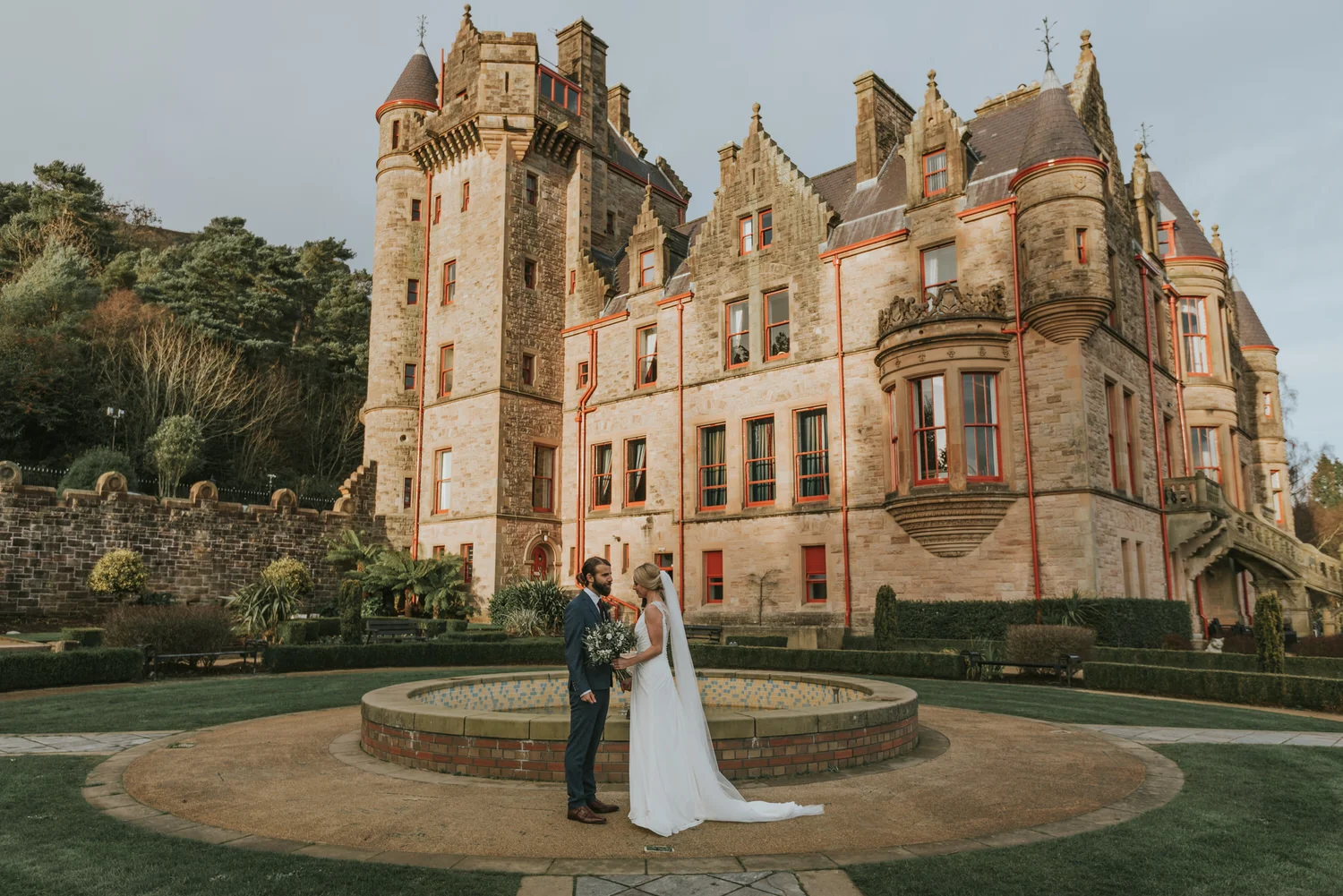 Bride and groom standing together by the fountain in the gardens in front of Belfast Castle