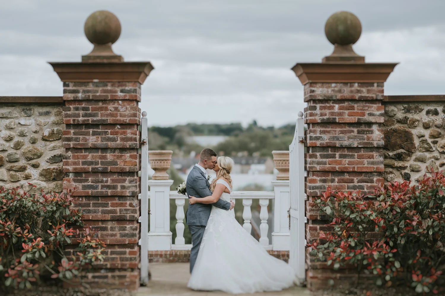 Bride and groom kissing between brick gate pillars with the river town of Lisburn behind them at Lagan Valley Island