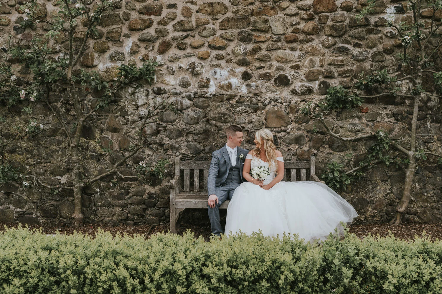 Bride and groom sitting together on a wooden bench in front of an old stone wall at Lagan Valley Island, Lisburn