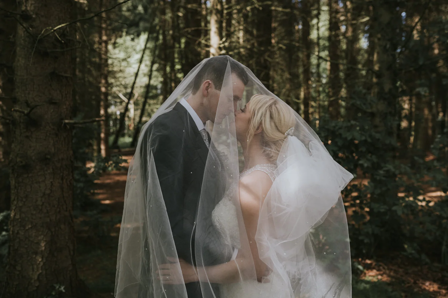 Bride and groom kissing beneath her veil among tall trees in woodland on their wedding day