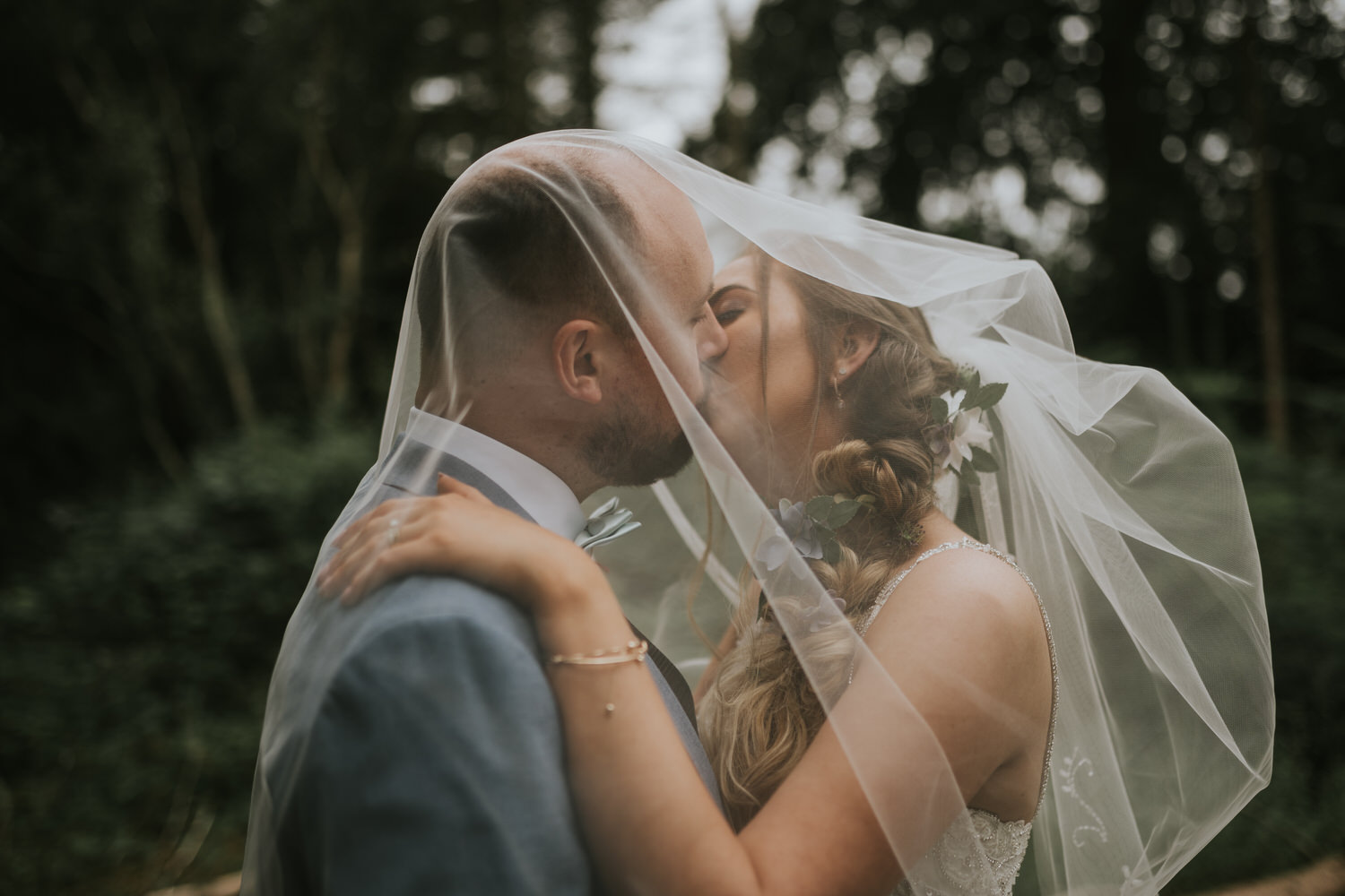 Bride and groom kissing beneath her veil among the trees on their wedding day