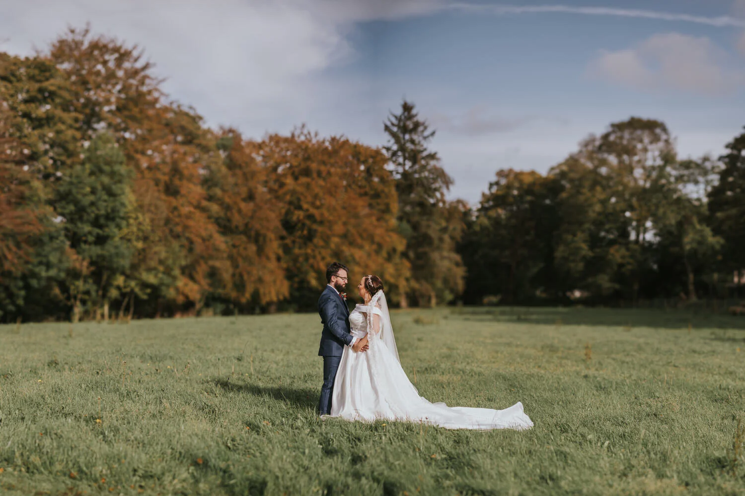 Bride and groom embracing in a field with autumn trees behind them at The Wool Tower