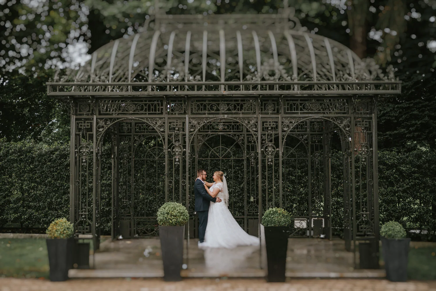 Bride and groom embracing in front of an ornate iron gazebo in the gardens at Millbrook Lodge