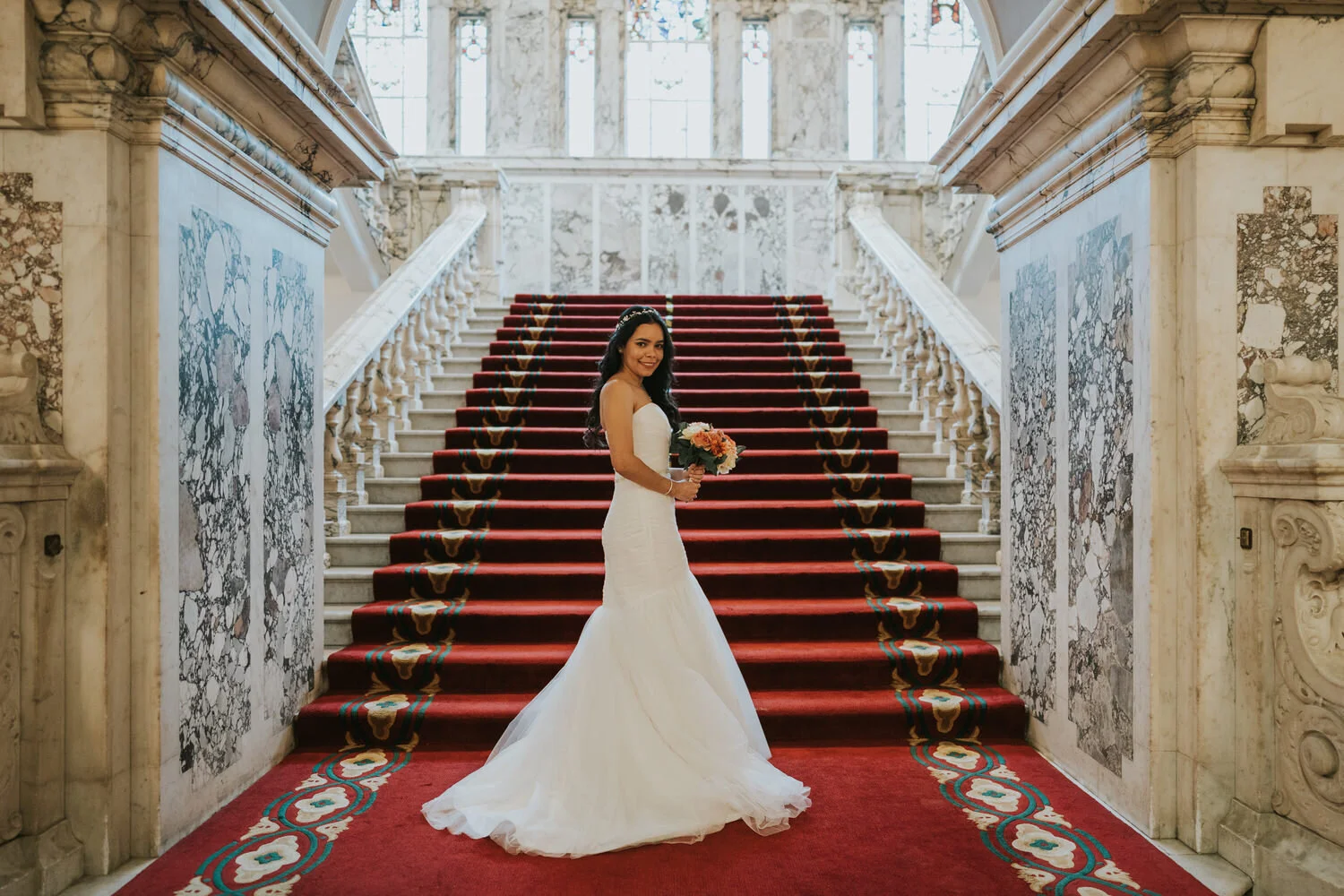 Bride in a white gown standing on the red carpeted marble staircase inside Belfast City Hall