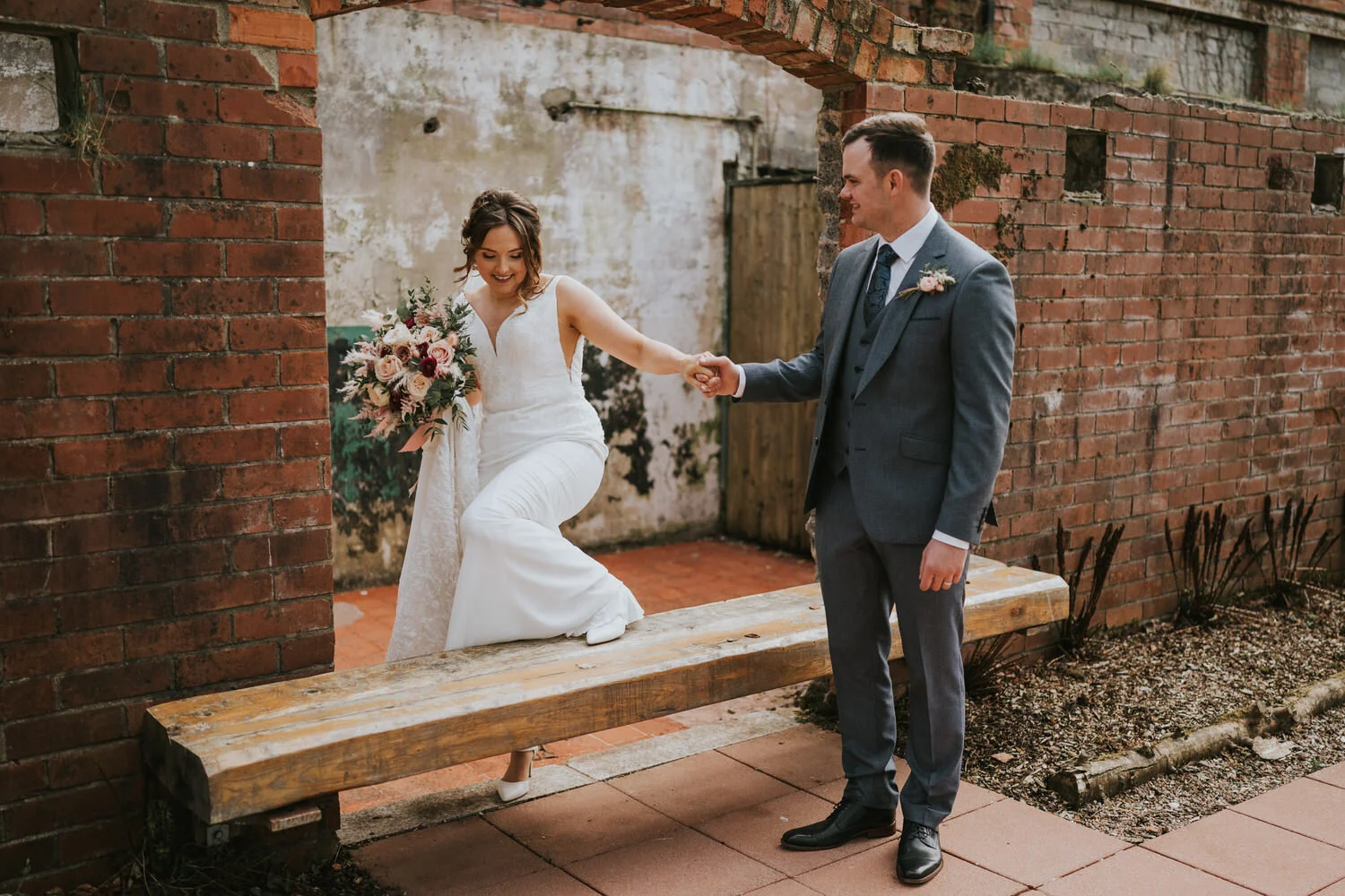 Bride stepping along a wooden bench, holding the groom's hand in front of a brick archway at The Wool Tower