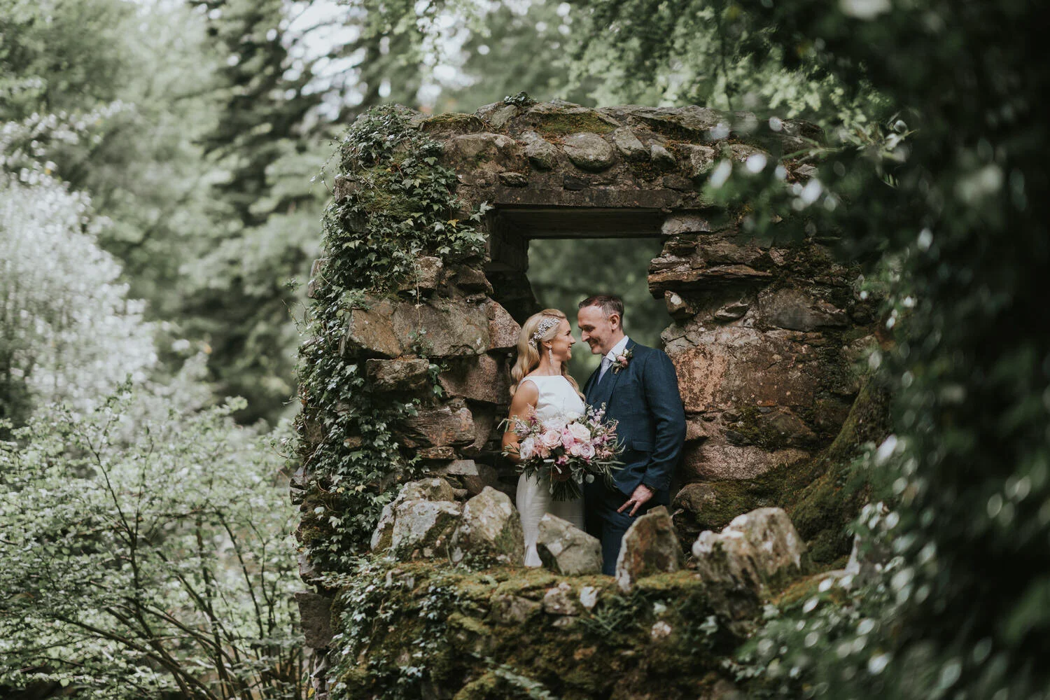 Bride and groom smiling at each other inside a moss covered stone ruin surrounded by woodland near the Slieve Donard hotel