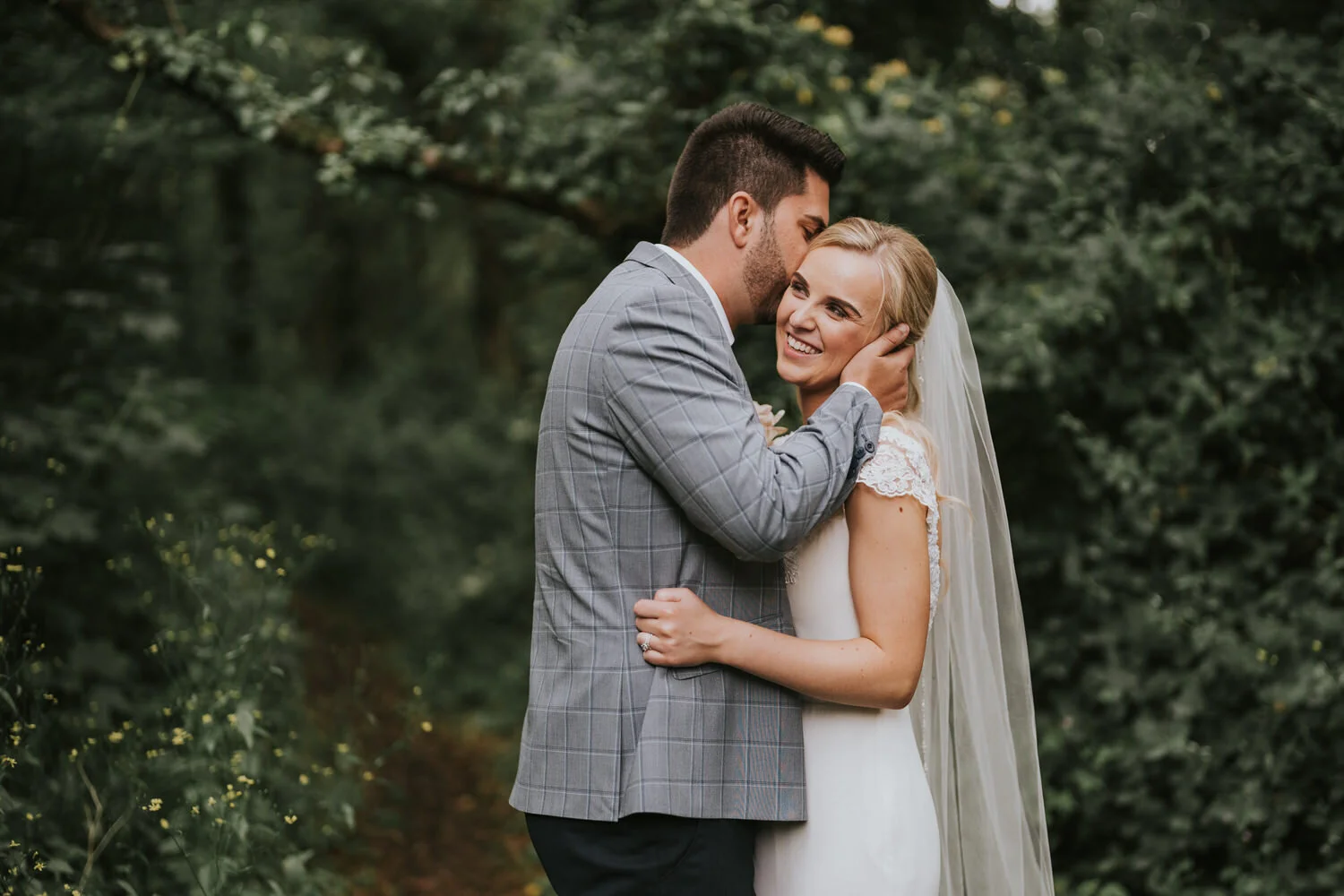 Groom kissing the bride on the cheek as she smiles, in the woodland at Lusty Beg Island
