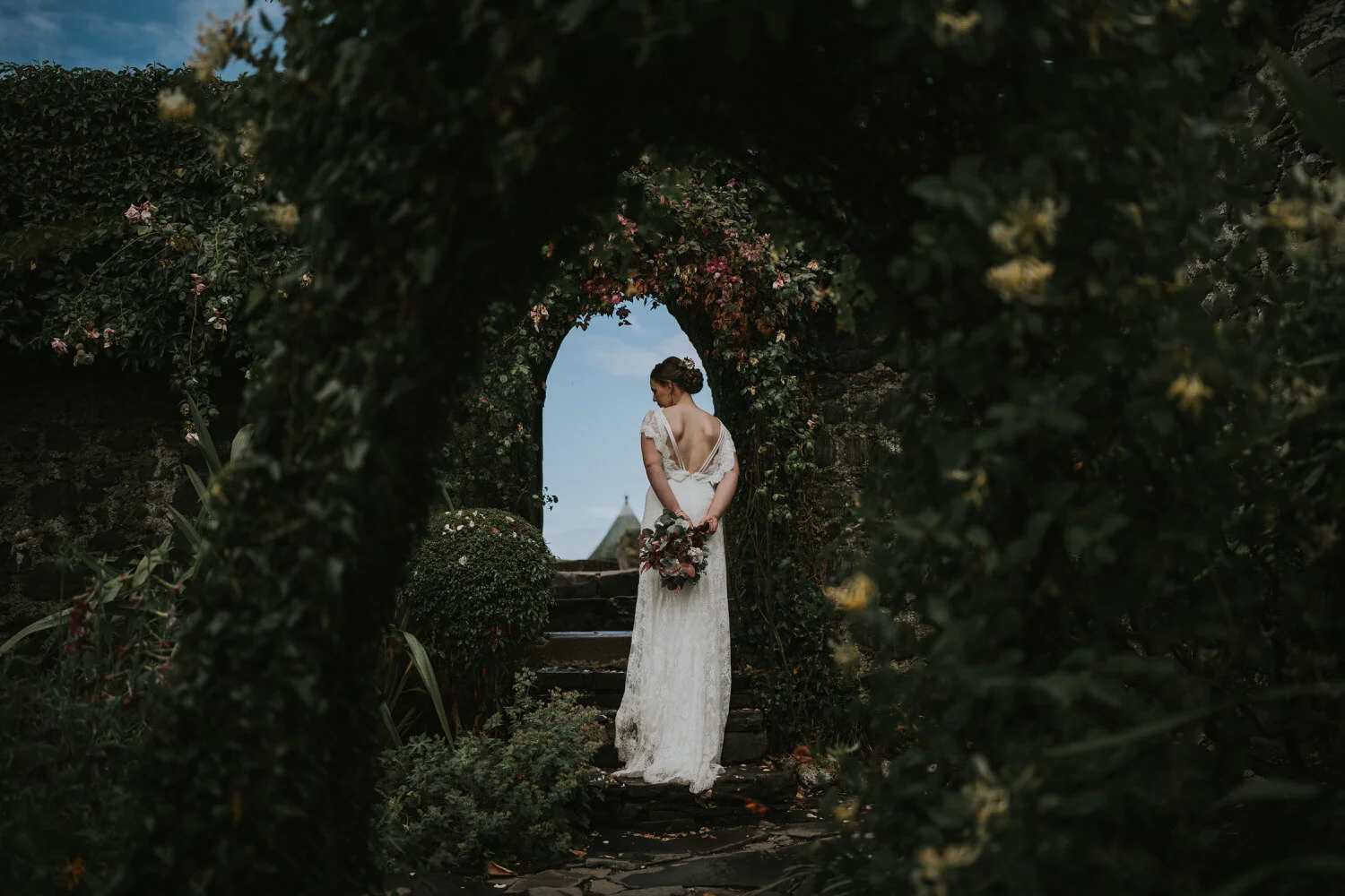 Bride in a lace wedding dress holding her bouquet behind her back, framed by an arch of foliage at Ballygally Castle