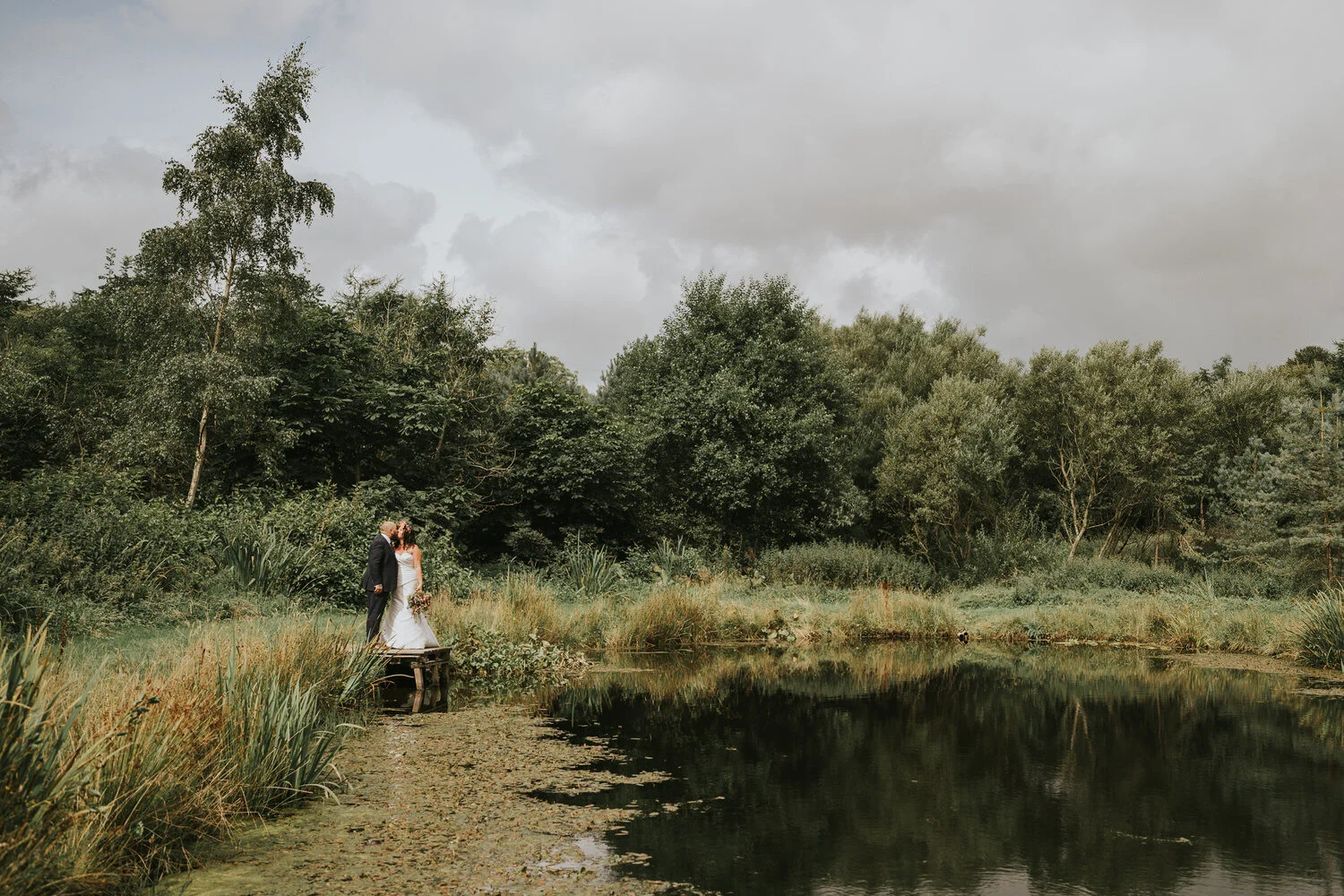 Bride and groom embracing beside the lake in the gardens at Breckenhill, Ballyclare