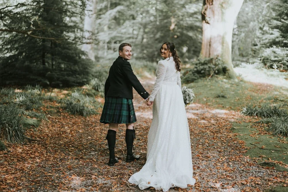 Bride and groom holding hands and looking back over their shoulders on a leaf covered woodland path near the Slieve Donard hotel