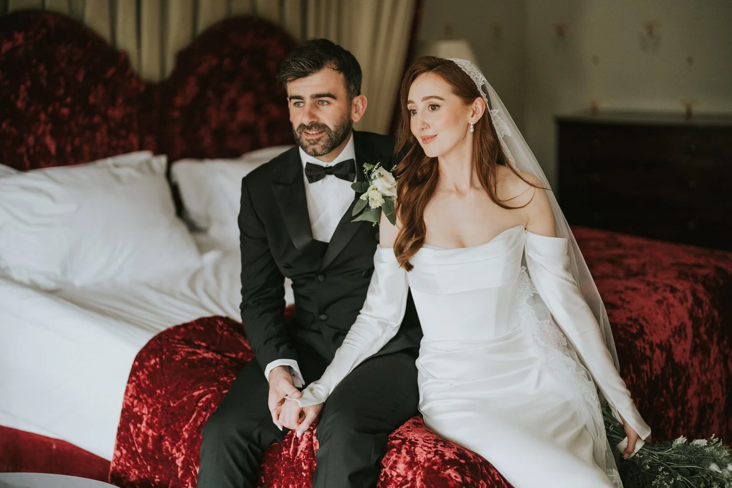 Bride and groom holding hands on a bed with a red velvet headboard in a bedroom at Beech Hill Country House