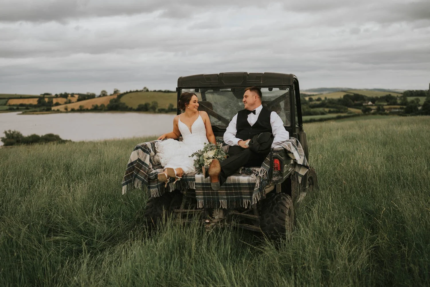 Bride and groom sitting together in a small vehicle in a grassy field overlooking the water near Tullyveery House