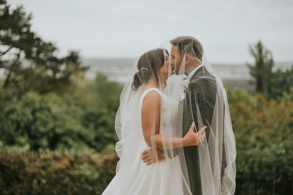 Maria and Sean kissing under her veil in the gardens at Belfast Castle with the lough behind