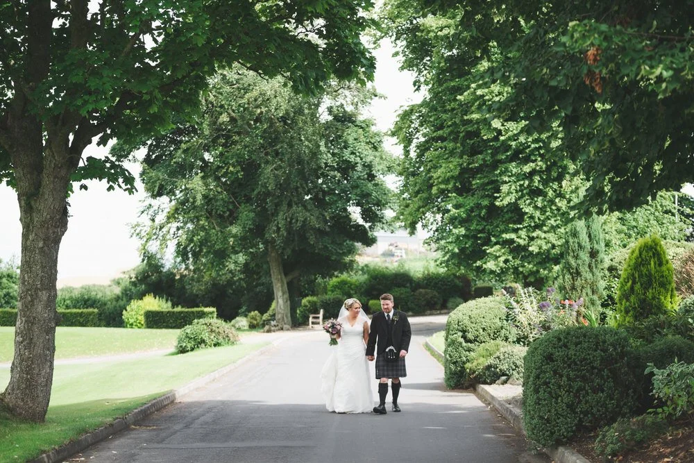 Bride and groom in a kilt walking arm in arm along a tree lined drive in the grounds at Edenmore House, Magheralin